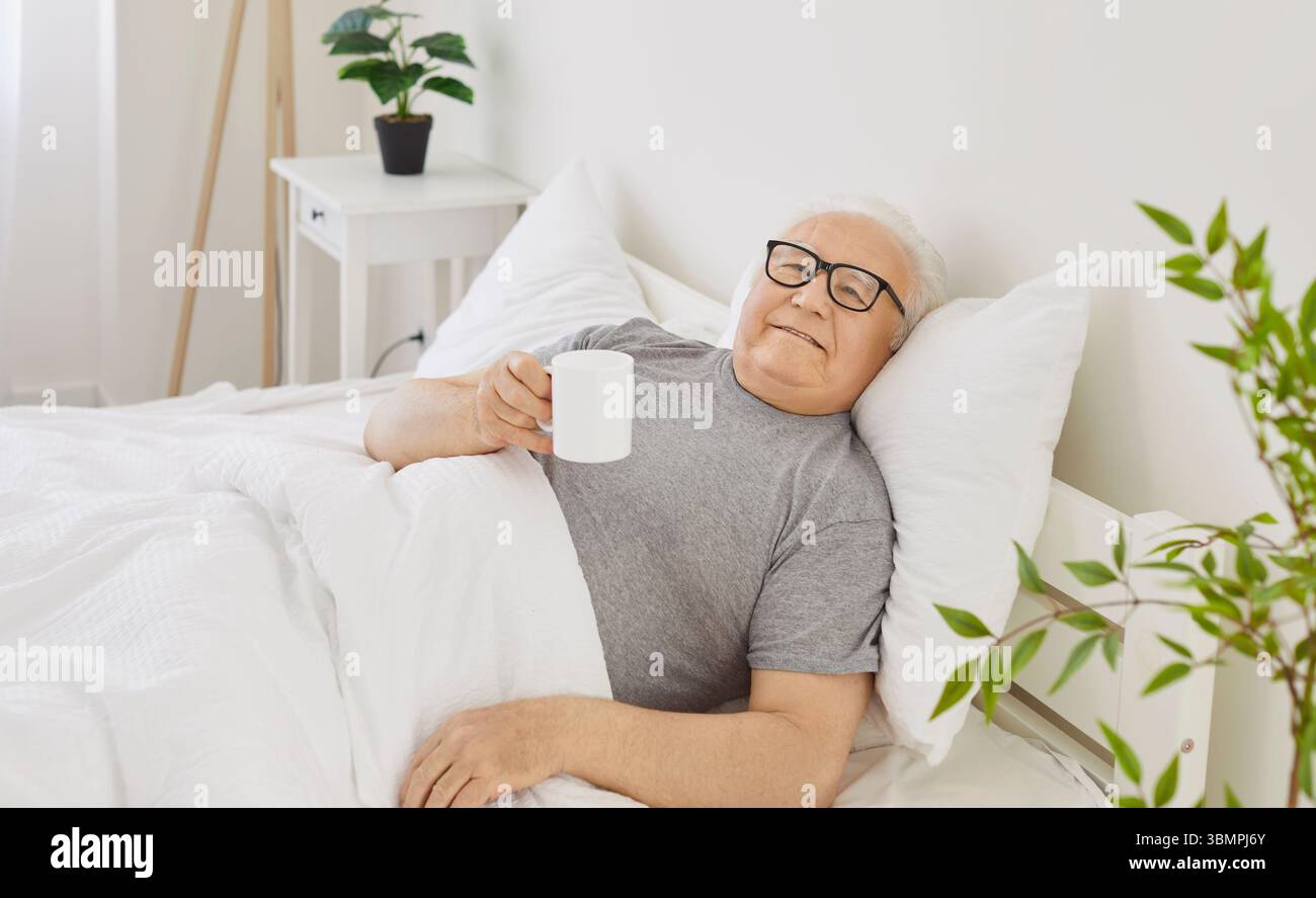 Homme âgé senior allongé sur le lit dans la chambre tenant une tasse de café ou de thé appréciant bonjour Banque D'Images