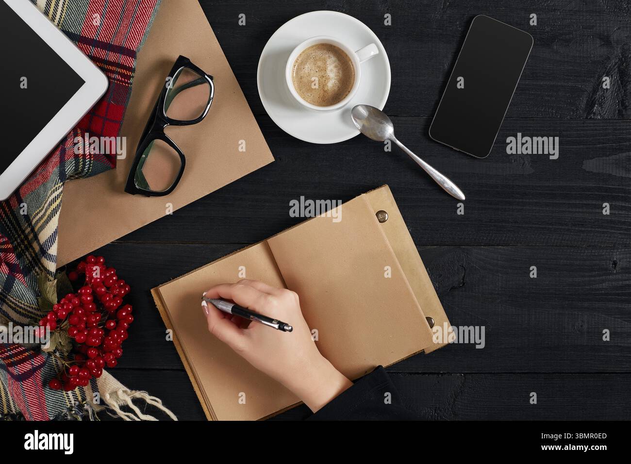 Jeune femme droite écrivant sur un carnet vierge sur une table en bois avec une tasse de café, un smartphone et des lunettes à côté dans l'heure du matin. Vue de dessus. Encore la vie. Banque D'Images