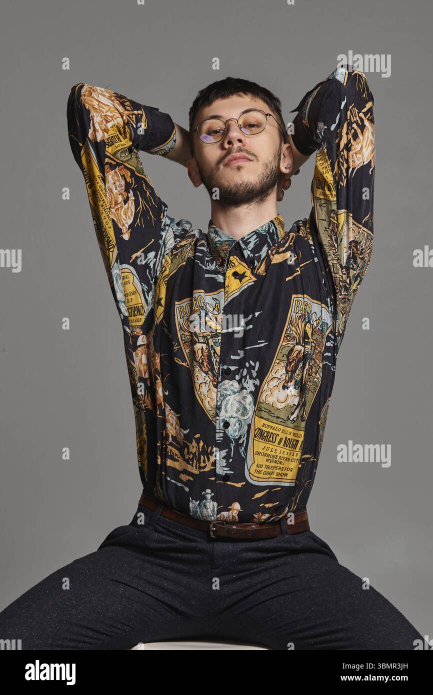 Portrait en gros plan d'un beau brun mâle dans des lunettes, porté dans une chemise colorée, regardant l'appareil photo tout en étant assis sur une chaise de bar dans le studio à nouveau Banque D'Images
