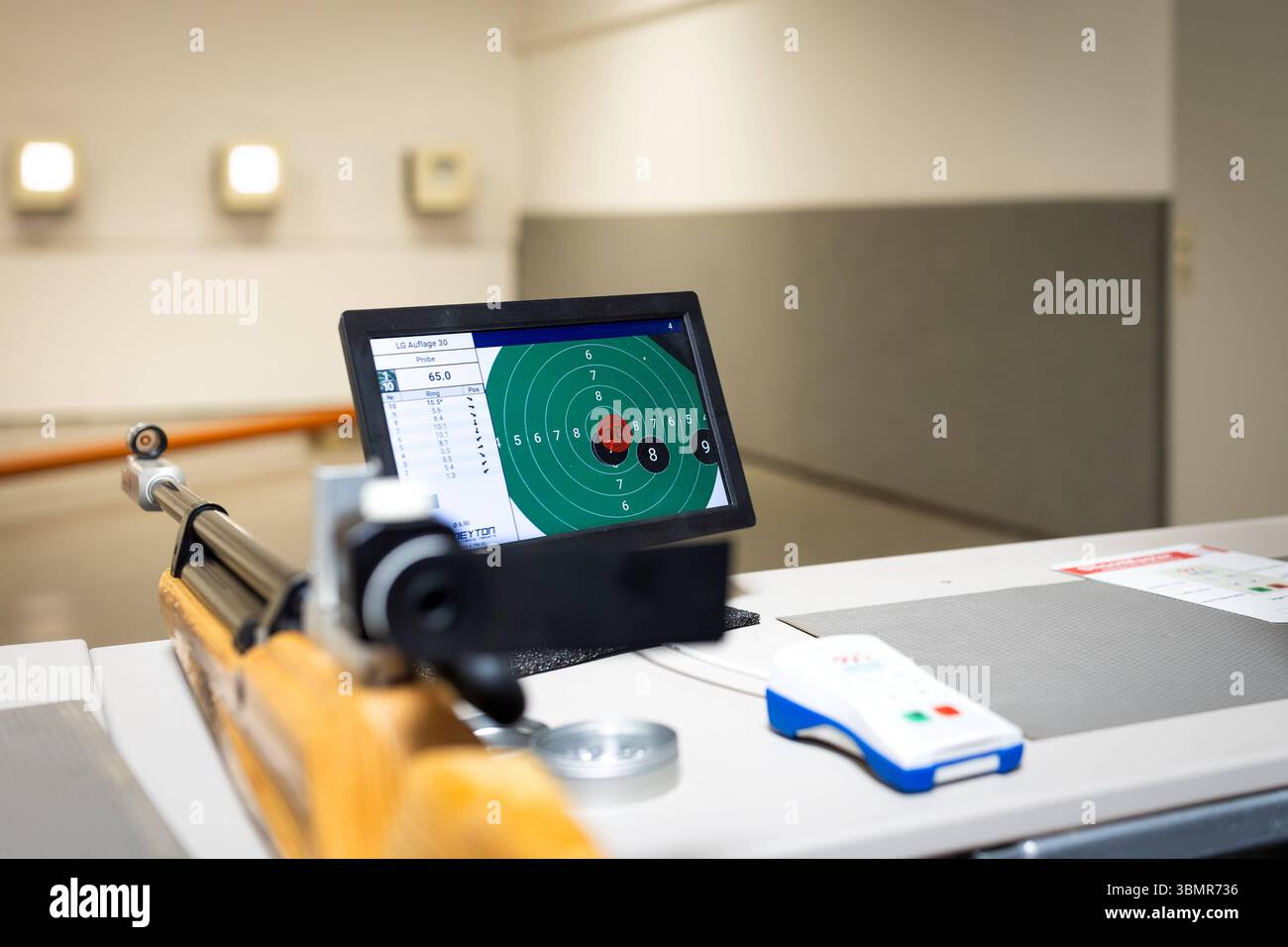 Landkreis Osnabrück, Deutschland 25. Juni 2025 : Im Bild : Ein Schießstand wird aus der Perspektive des Schützen gezeigt, mit einem klassischen Holzschaft-Luftgewehr im Vordergrund, das auf einem Tisch aufliegt. Der Fokus liegt auf einem LCD-Monitor im Mittelpunkt des Bildes, der eine digitale Zielscheibe und die Ergebnisse des Luftgewehr LG Auflage 30 Schießens anzeigt. Dies demonstriert die Integration moderner elektronischer Zielerfassungssysteme in den traditionellen Schießsport. Rechts neben dem Monitor befindet sich eine weiße Fernbedienung mit blauen und roten Akzenten, die zur Steuerung Banque D'Images