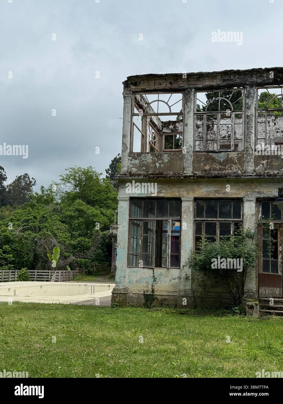 La photo montre un bâtiment de deux étages détérioré avec des fenêtres endommagées, un cadre exposé et des murs délavés. La structure est partiellement dépassée par ve Banque D'Images