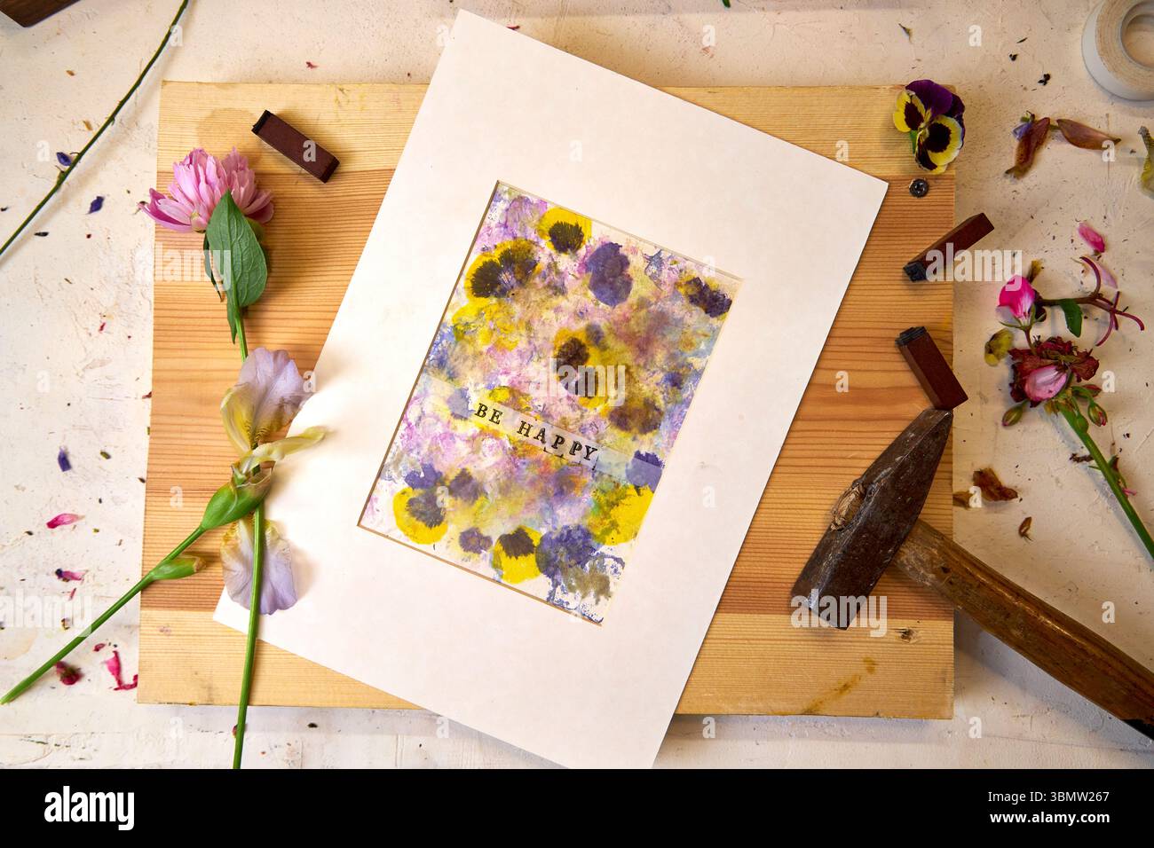 Imprimé botanique fait à la main avec texte « Be Happy », mat et prêt à être affiché – pigments floraux, marteau, fleurs et timbres dans un espace de travail créatif Banque D'Images