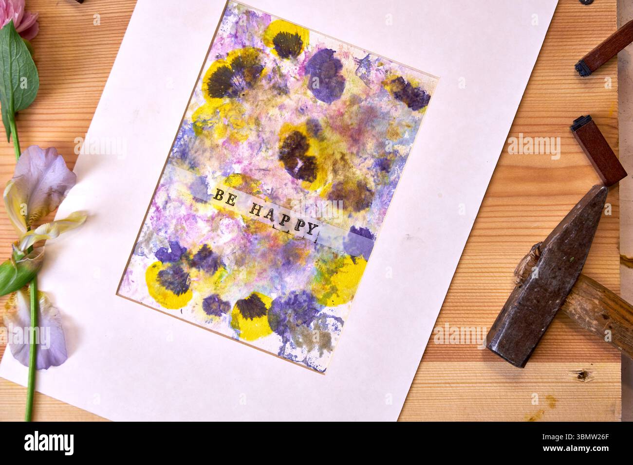 Imprimé botanique fait à la main avec texte « Be Happy », mat et prêt à être affiché – pigments floraux, marteau, fleurs et timbres dans un espace de travail créatif Banque D'Images