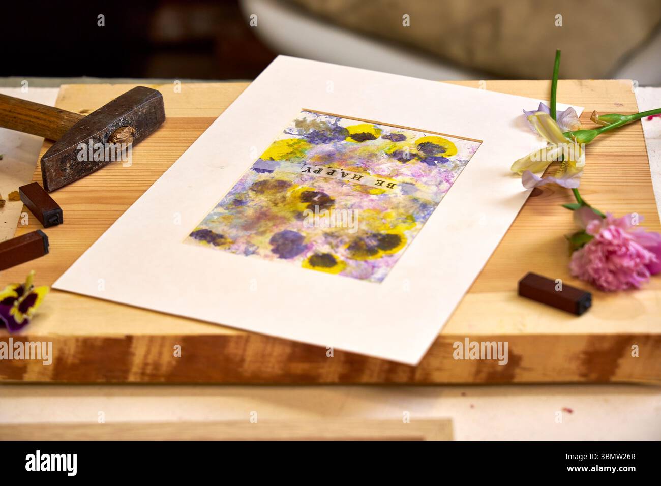 Imprimé botanique fait à la main avec texte « Be Happy », mat et prêt à être affiché – pigments floraux, marteau, fleurs et timbres dans un espace de travail créatif Banque D'Images