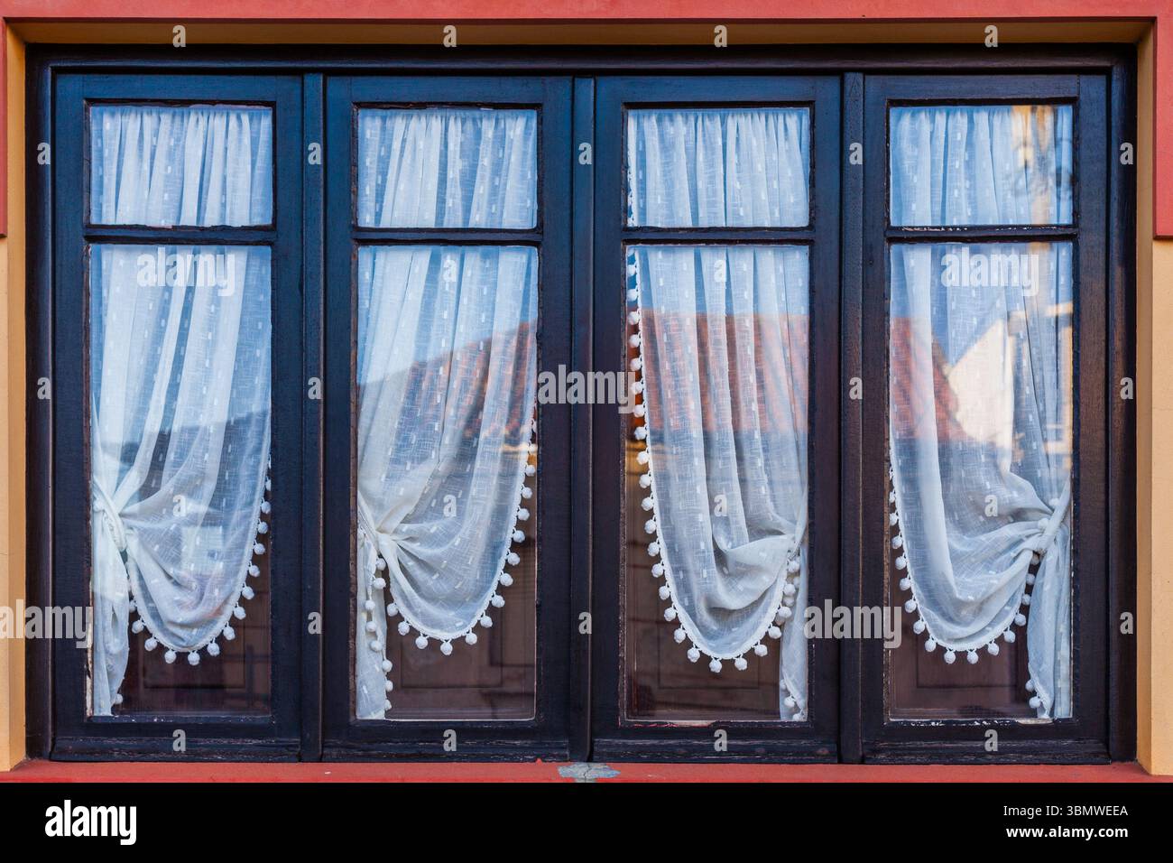 Une rangée de quatre fenêtres à cadre sombre avec des rideaux de dentelle décoratifs clairs et des reflets visibles offre un aperçu d'une maison européenne traditionnelle Banque D'Images