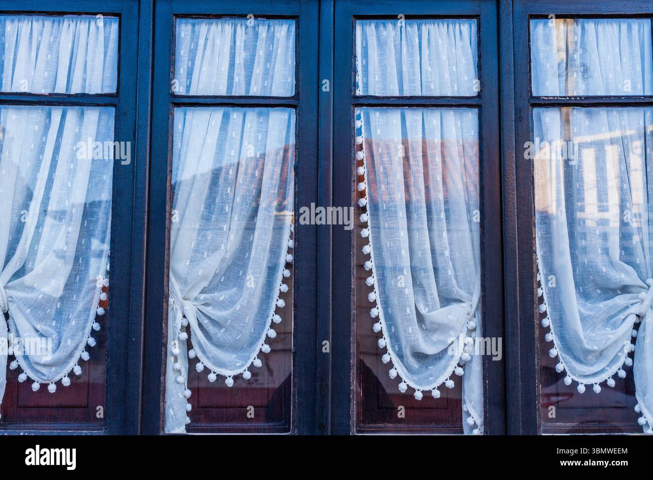 Une rangée de quatre fenêtres à cadre sombre avec des rideaux de dentelle décoratifs clairs et des reflets visibles offre un aperçu d'une maison européenne traditionnelle Banque D'Images