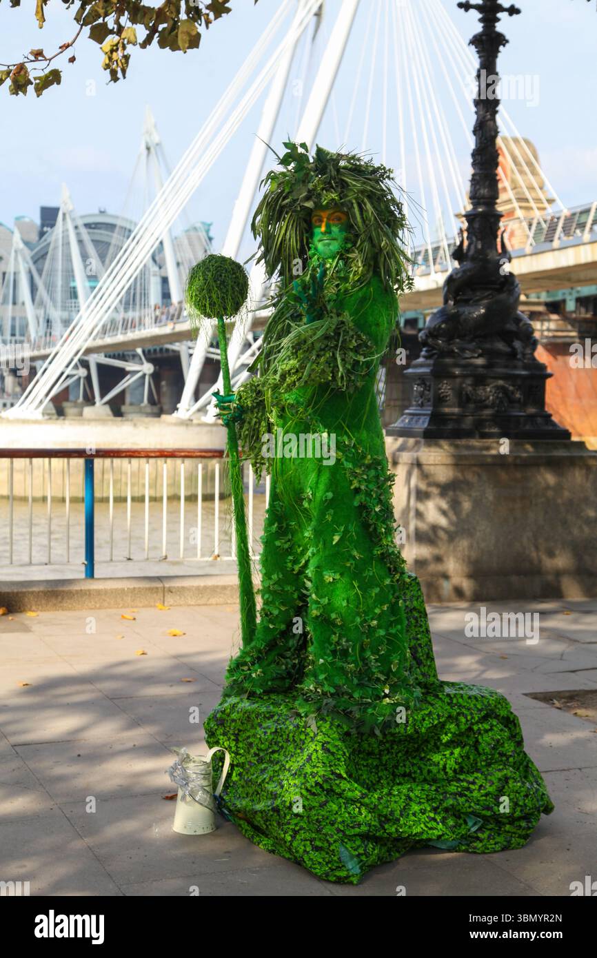 Artiste habillé comme une statue verte vivante avec le personnel près de Millennium Bridge sur South Bank, Londres - mêlant art, nature et vie urbaine. Banque D'Images