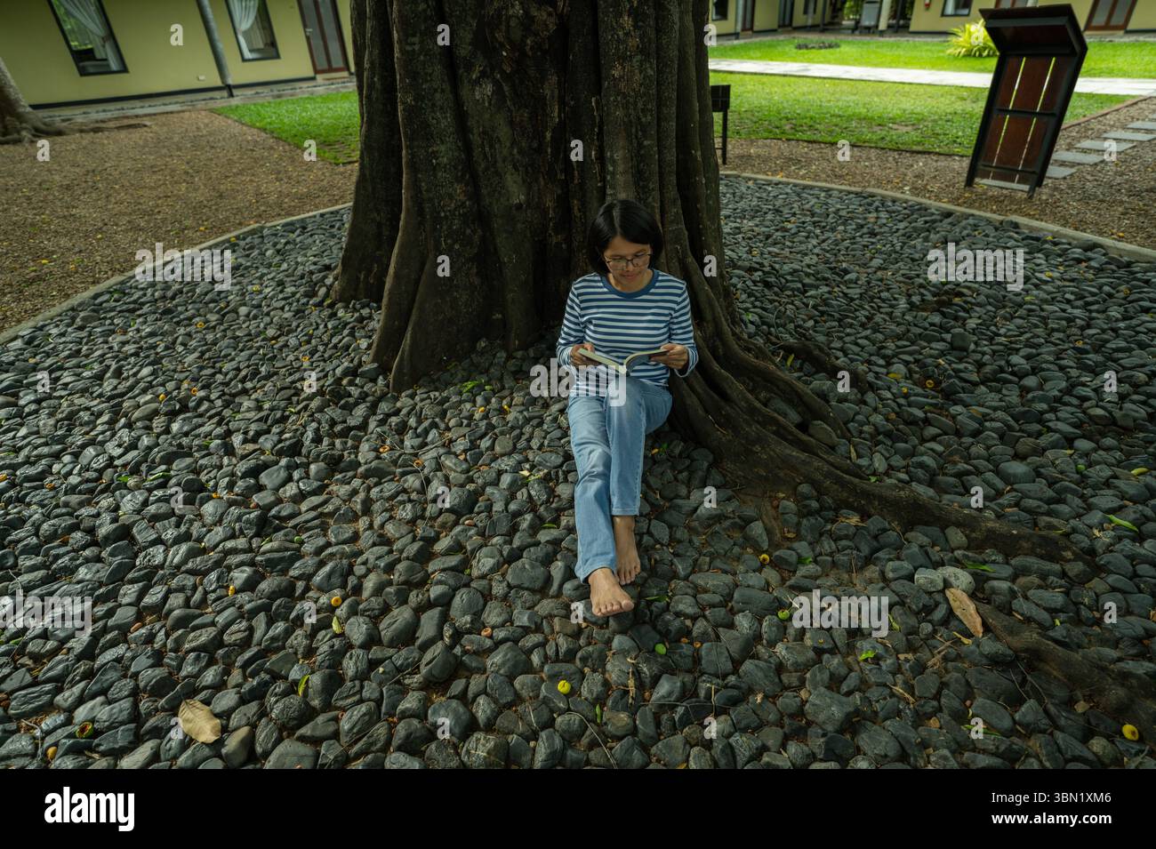 Femme thaïlandaise assise pieds nus lisant livre sous l'arbre au complexe hôtelier de luxe. Retraite de vacances paisible. Détox numérique et relaxation de bien-être attentif. Banque D'Images