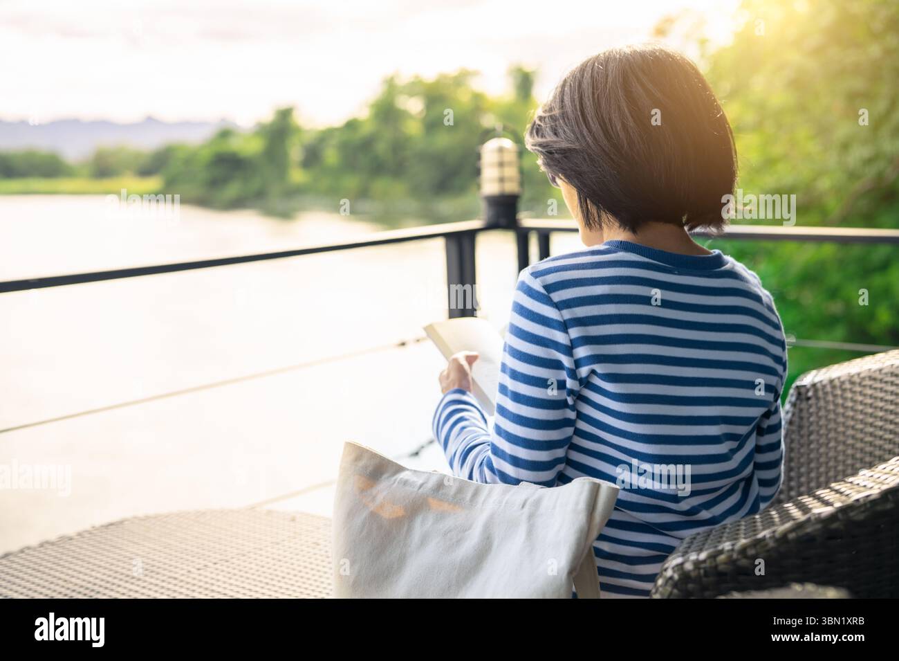 Femme assise sur la chaise lisant un livre sur le balcon surplombant la rivière pendant l'heure dorée matin. Soins personnels, détox numérique et vie lente consciente. EMO Banque D'Images