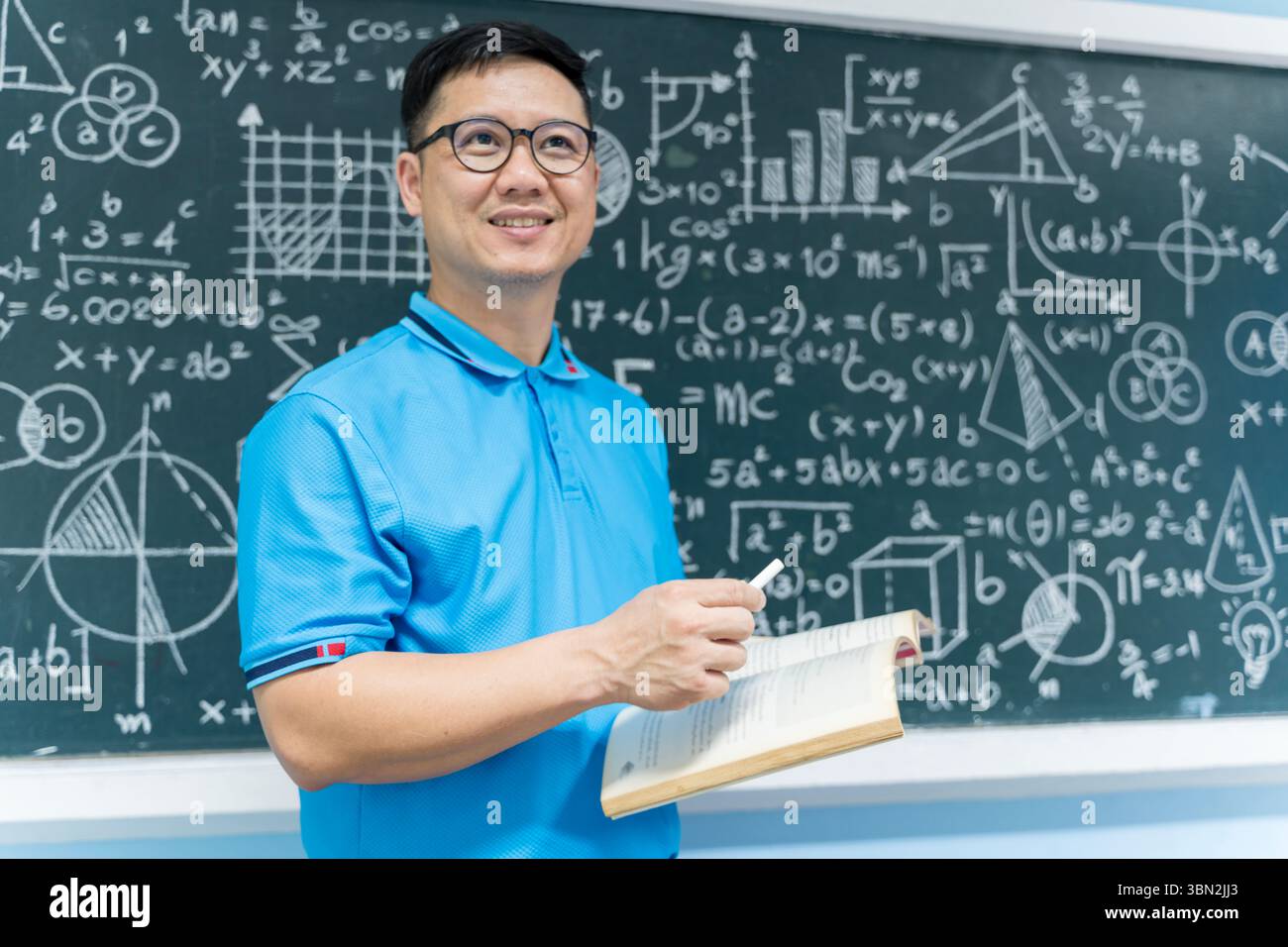 Portrait d'un professeur asiatique souriant tenant un livre ouvert et de la craie devant un tableau noir avec des formules mathématiques. Idéal pour le contenu lié à educ Banque D'Images