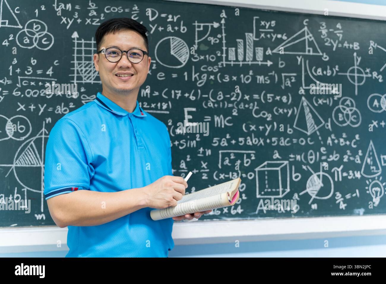 Portrait d'un enseignant asiatique tenant un livre ouvert et de la craie devant un tableau rempli de formules mathématiques. Idéal pour le contenu sur educ Banque D'Images