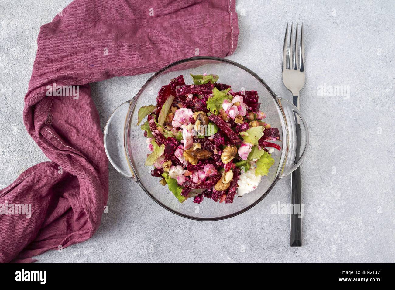 Salade de betteraves, feuilles vertes, fromage feta, noix dans une assiette en verre, une serviette Bordeaux et une fourchette reposent sur une table grise Banque D'Images