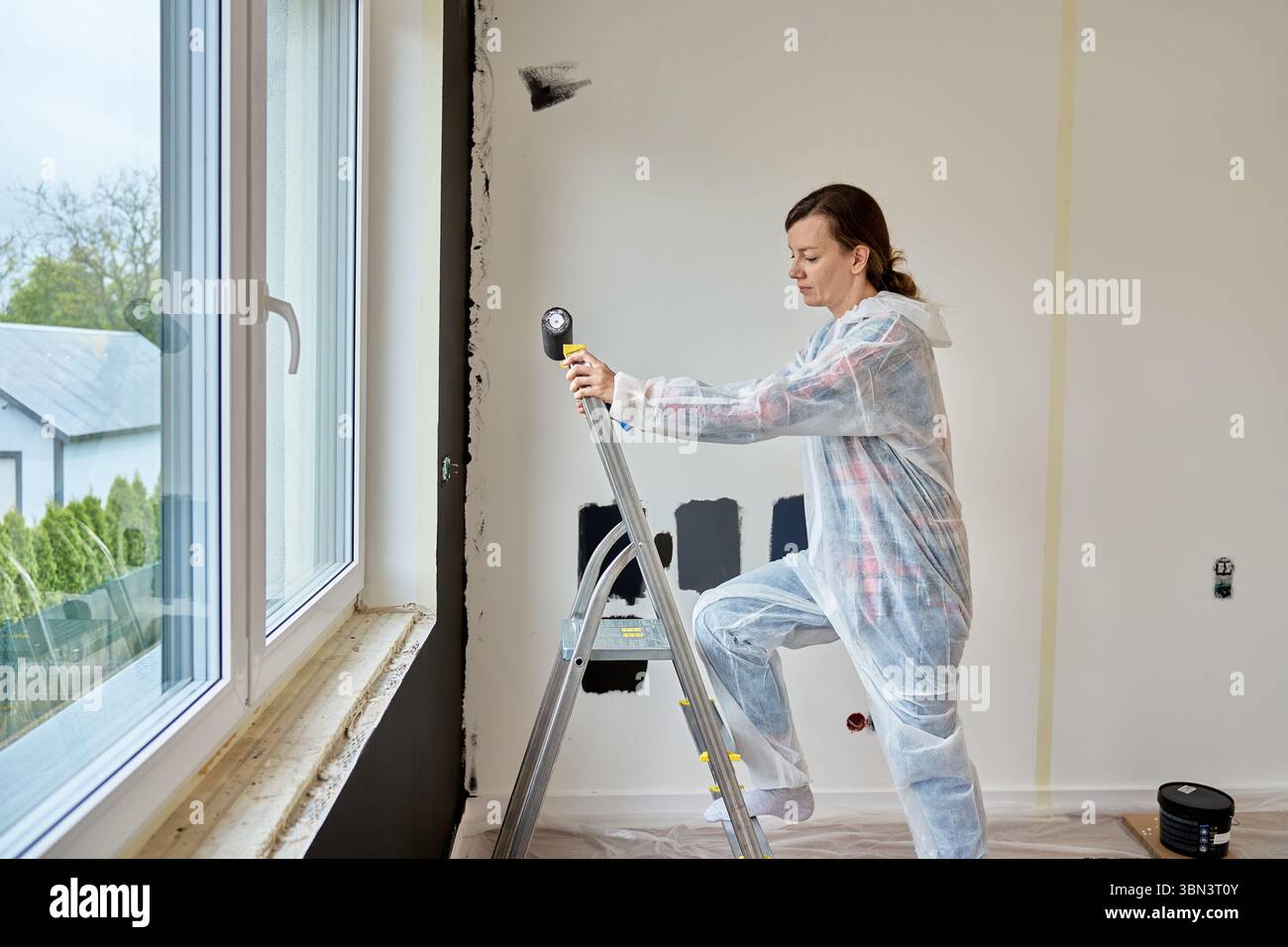 Femme dans l'échelle de montée d'usure de protection avec rouleau de peinture dans la main près de la fenêtre, se préparant à peindre le mur d'accent noir. Rénovation de la maison dans un appartement moderne Banque D'Images