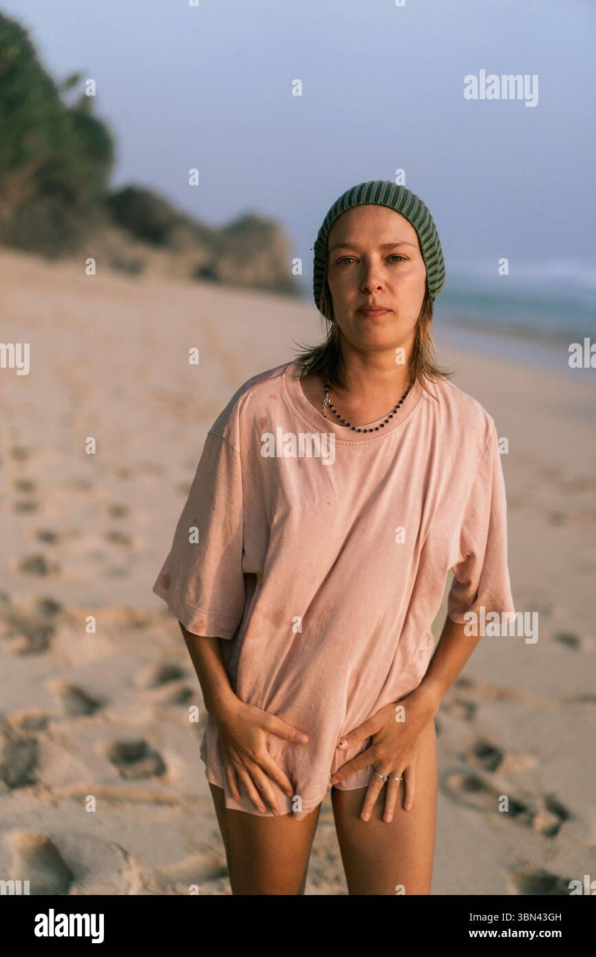 Jeune femme en chemise rose et bonnet vert se tient sur la plage au lever du soleil Banque D'Images