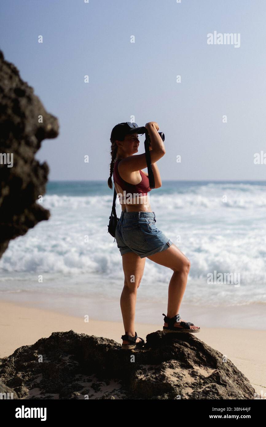 Femme voyageuse avec appareil photo prenant des photos de l'océan de rocher sur la plage Banque D'Images