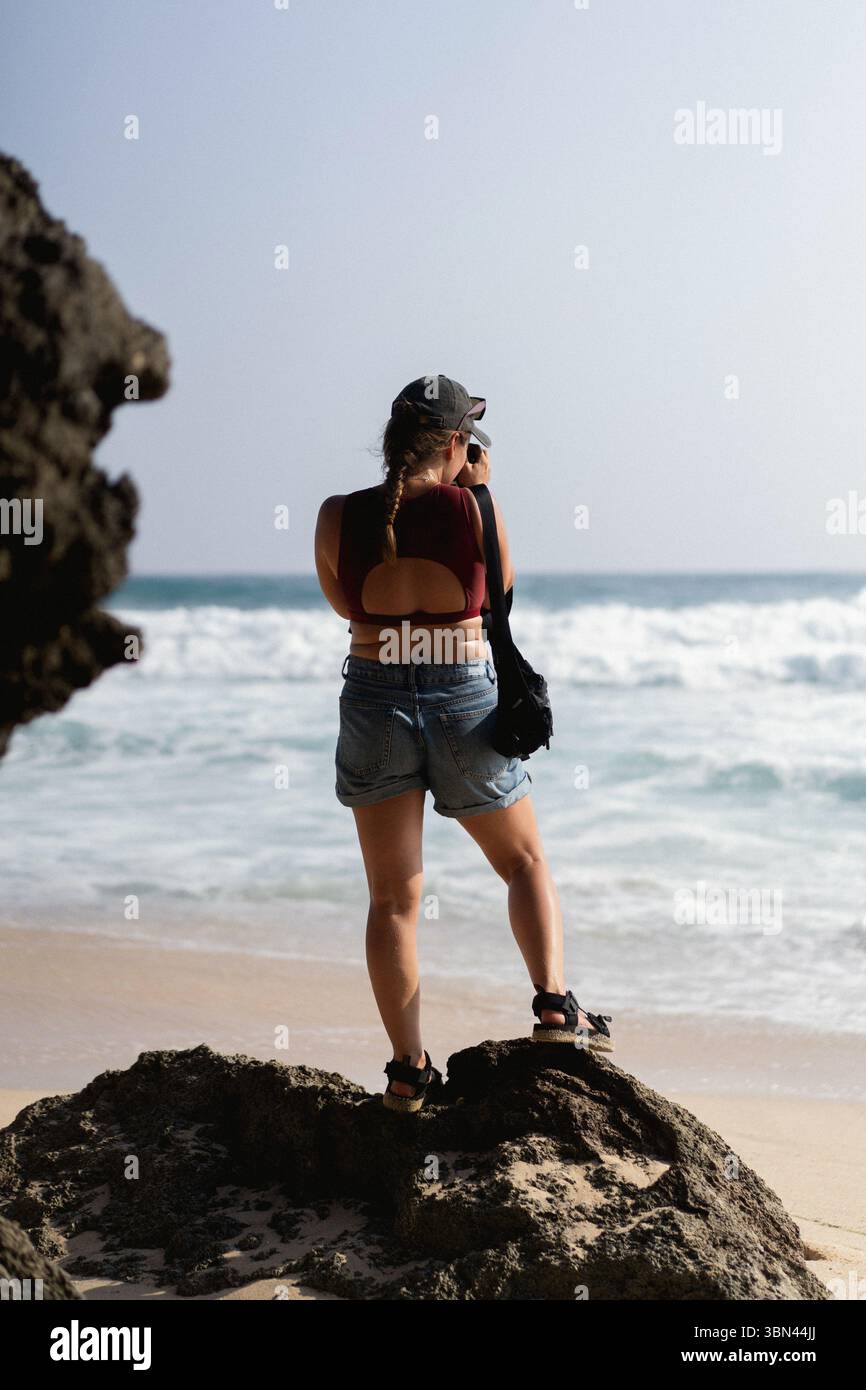 Femme voyageuse avec appareil photo prenant des photos de l'océan de rocher sur la plage Banque D'Images