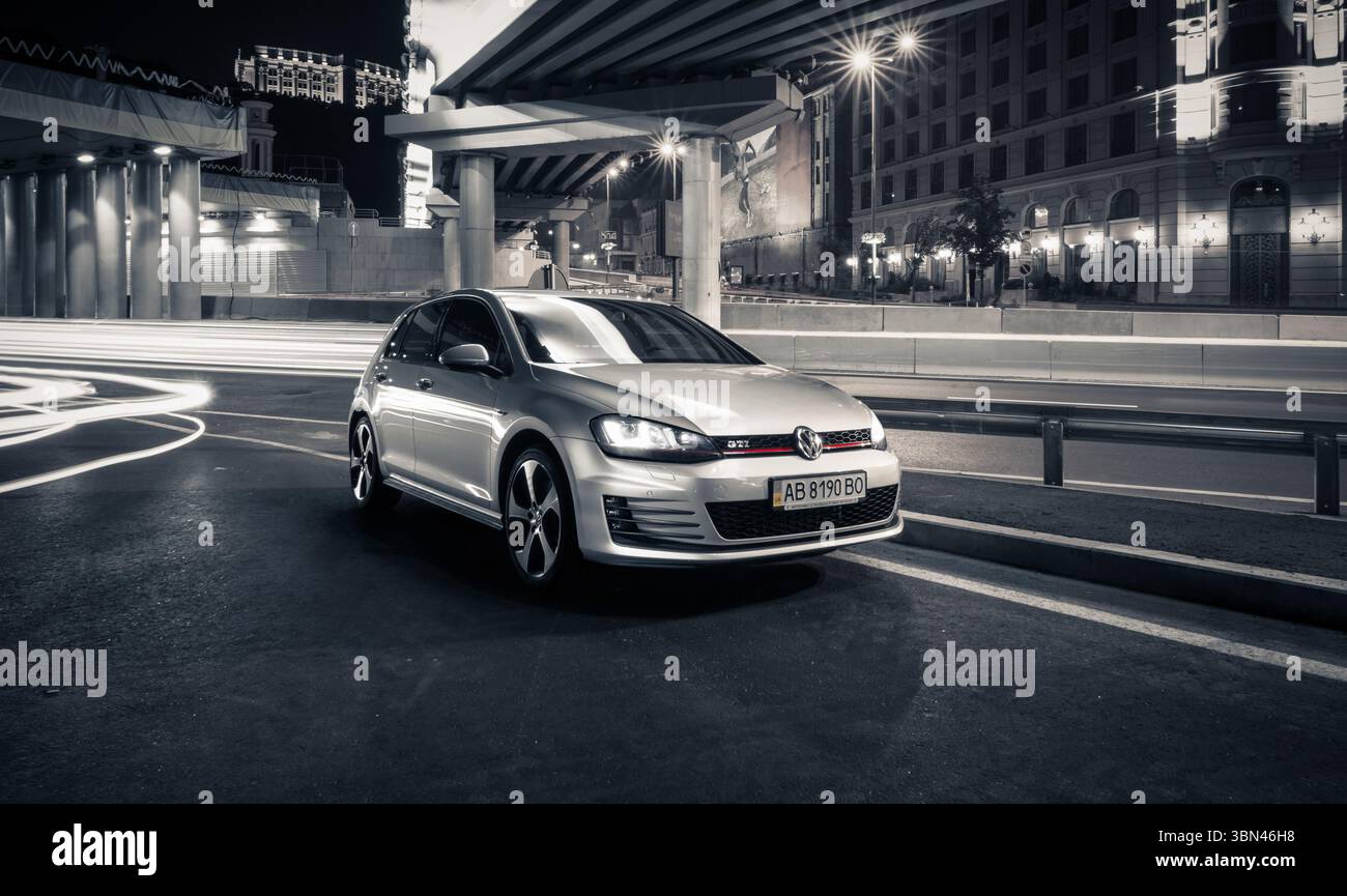 Silver Volkswagen Golf GTi sur la rue de la ville de nuit. Vue de trois quarts de face de la VW à hayon - image longue exposition avec traînées de lumière. Banque D'Images