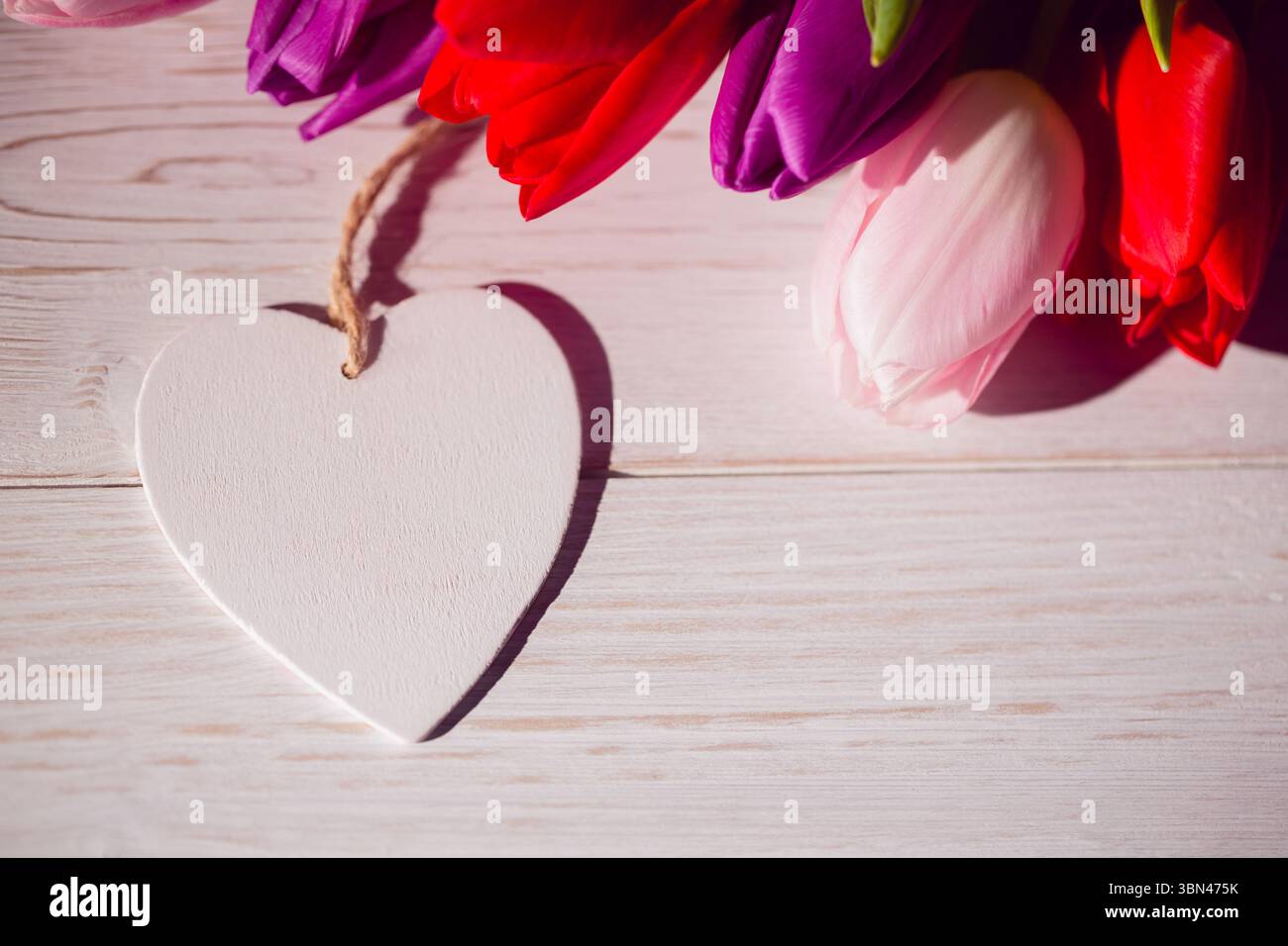 L'ornement de coeur en bois blanc de conception plate repose sur une table avec un bouquet de tulipes rouge, rose et violet Banque D'Images