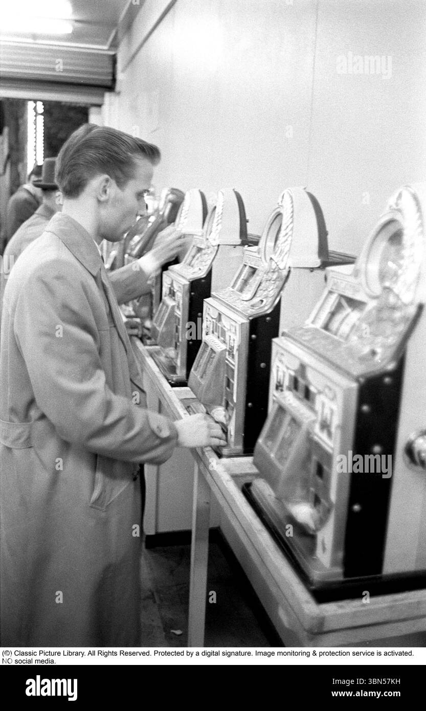 Aux machines à sous 1960. Les gens photographiés aux machines à sous. Une machine de jeu où vous mettez dans une pièce de monnaie et tirez un levier qui commence la rotation de trois roues. Une fois qu'ils s'arrêtent et que le jeu de roues montre le même symbole, vous gagnez. Conard ref 4251 Banque D'Images