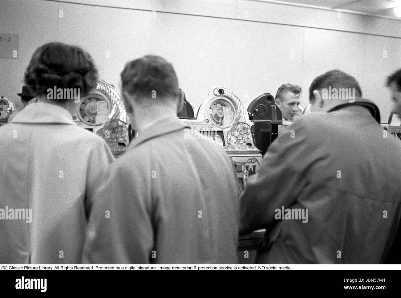 Aux machines à sous 1960. Les gens photographiés aux machines à sous. Une machine de jeu où vous mettez dans une pièce de monnaie et tirez un levier qui commence la rotation de trois roues. Une fois qu'ils s'arrêtent et que le jeu de roues montre le même symbole, vous gagnez. Conard ref 4251 Banque D'Images