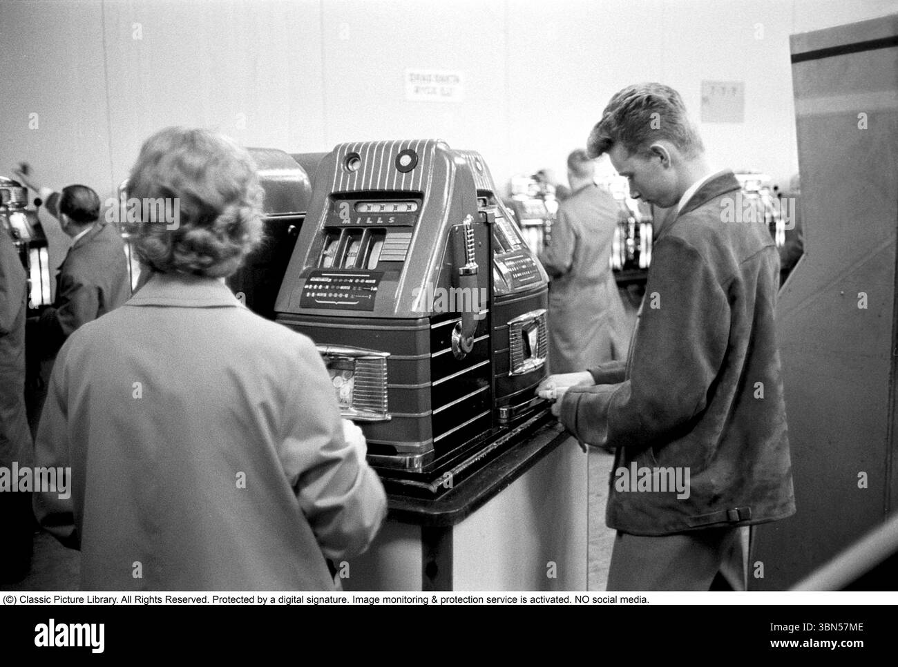 Aux machines à sous 1960. Les gens photographiés aux machines à sous. Une machine de jeu où vous mettez dans une pièce de monnaie et tirez un levier qui commence la rotation de trois roues. Une fois qu'ils s'arrêtent et que le jeu de roues montre le même symbole, vous gagnez. Conard ref 4251 Banque D'Images