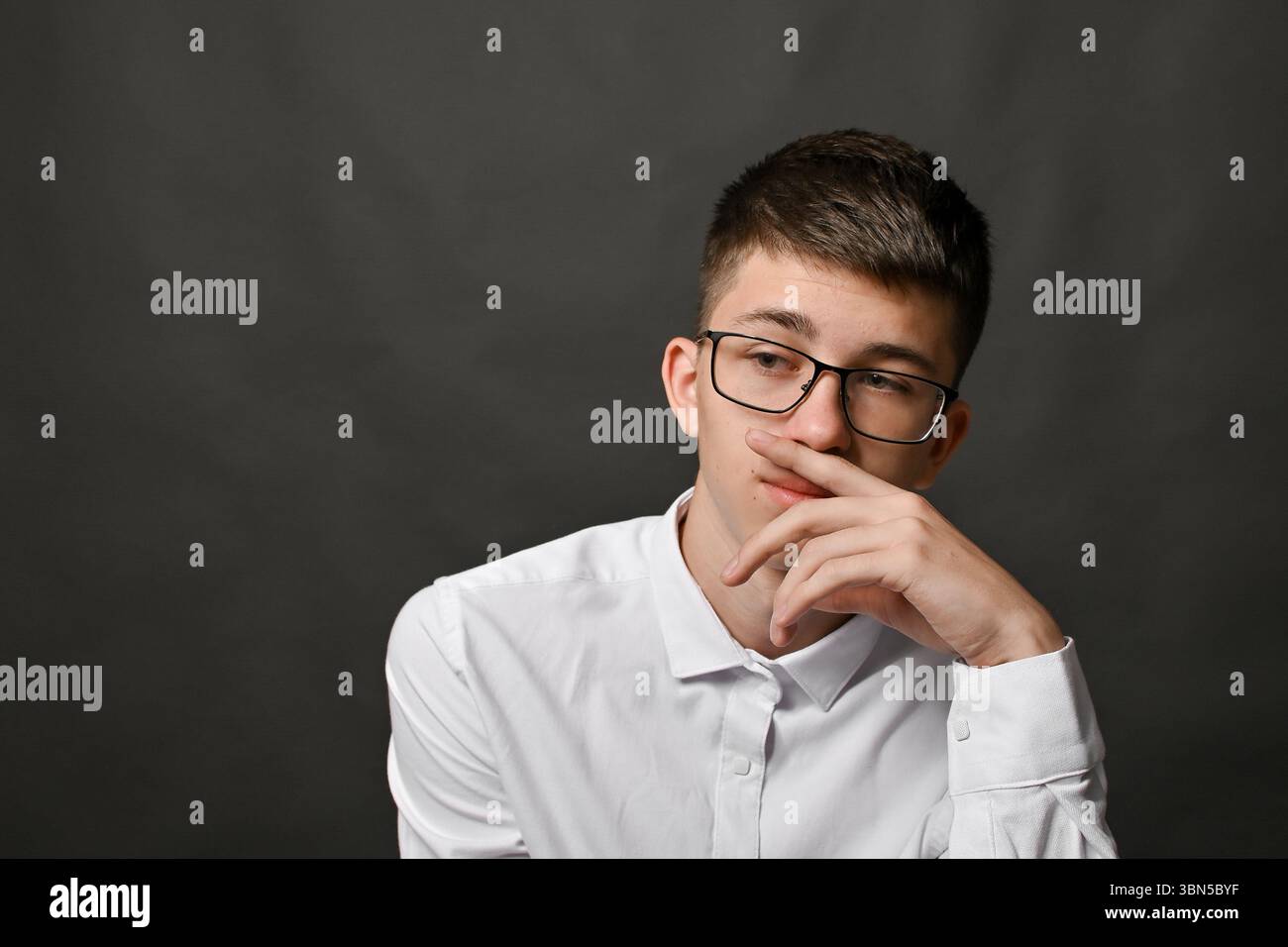 Jeune homme réfléchi dans des lunettes couvre la bouche avec la main sur fond gris de studio. L'expression pensive montre une concentration profonde et un mépris tranquille Banque D'Images