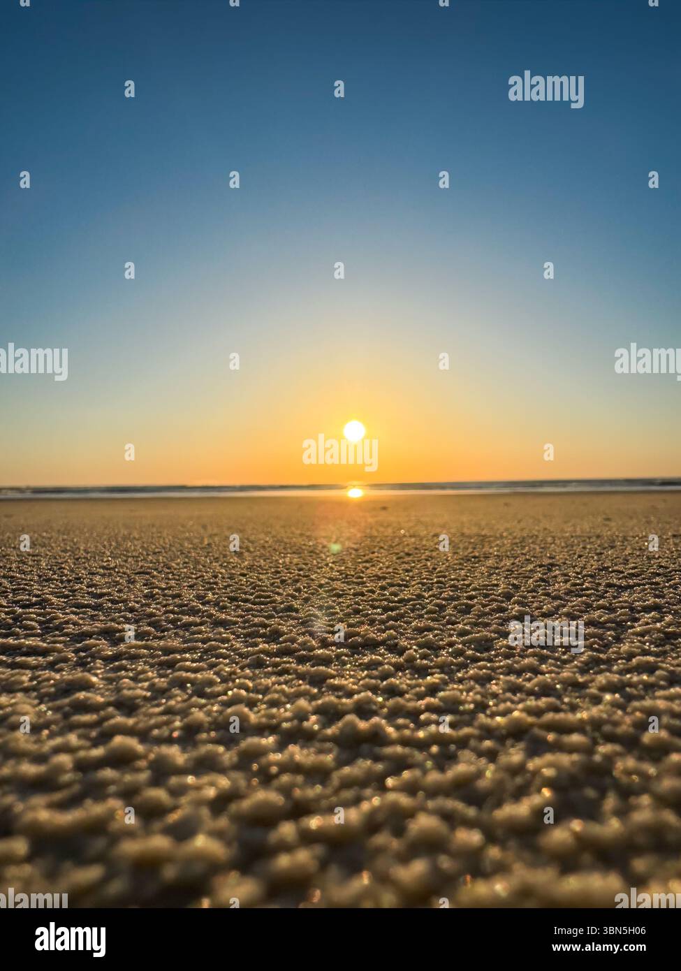 Une vue en bas angle du soleil levant sur une plage paisible. La lumière du matin scintille sur le sable texturé, capturant la beauté de la nature au lever du soleil. Banque D'Images