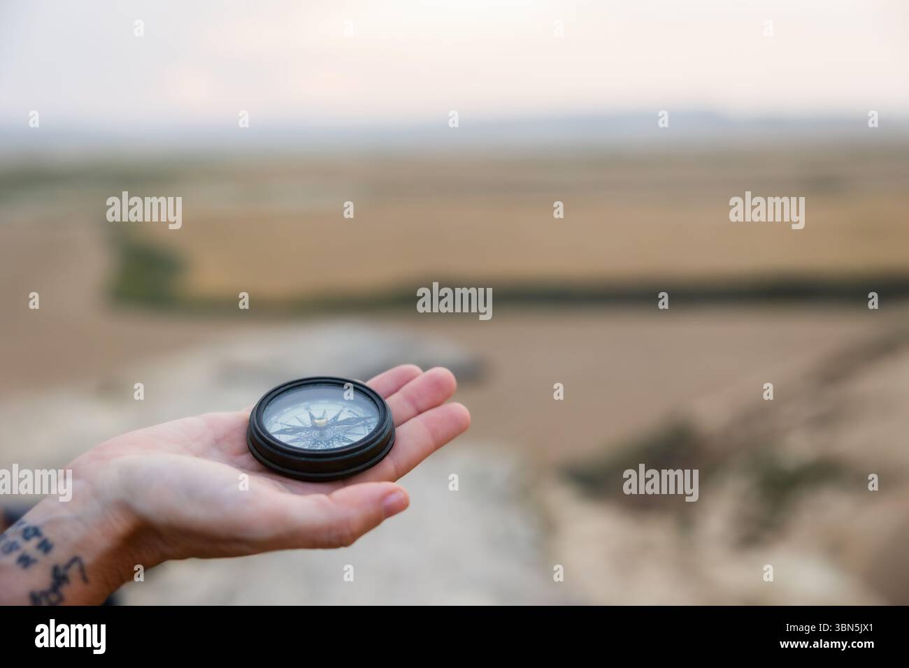 Une vue rapprochée d'une boussole reposant dans une main ouverte, avec un tatouage visible sur le poignet et un vaste paysage agricole légèrement flou au loin Banque D'Images