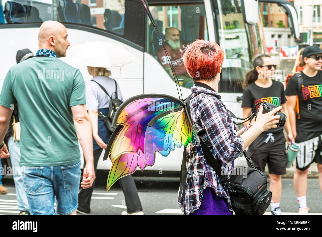 Femme aux cheveux rouges vibrants avec des ailes colorées, caméra à la main, regardant en arrière un vieux chauffeur de bus. Un curieux moment urbain. 28 juin 2025 Banque D'Images