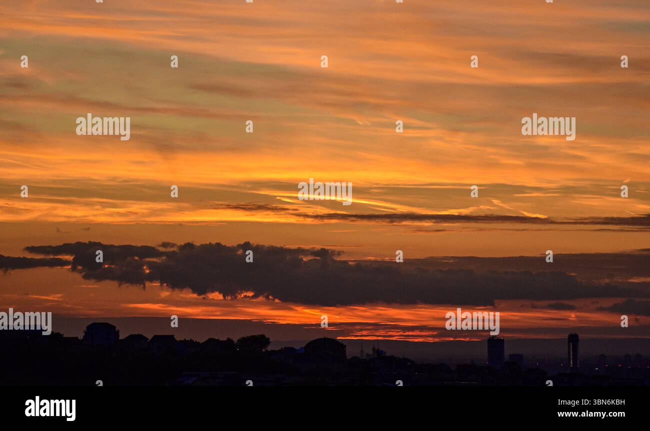 Le soleil couchant illumine le ciel dans des tons oranges tandis que les silhouettes des maisons et des bâtiments créent un paysage urbain spectaculaire. Banque D'Images