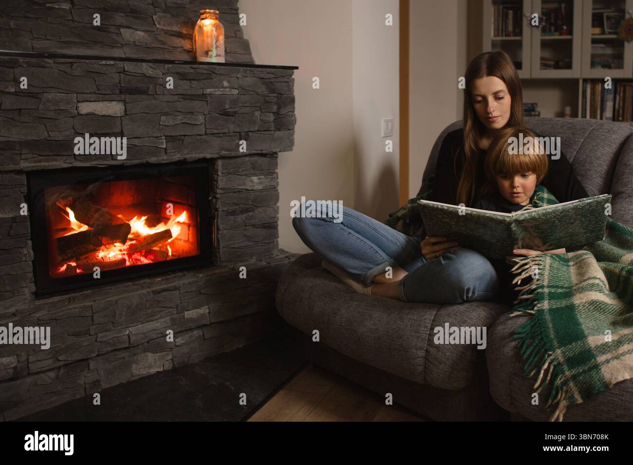 Mère et fils lisent un livre sur un canapé près de la cheminée dans un intérieur confortable. Amour, confort et famille ensemble dans une maison rustique moderne. Banque D'Images