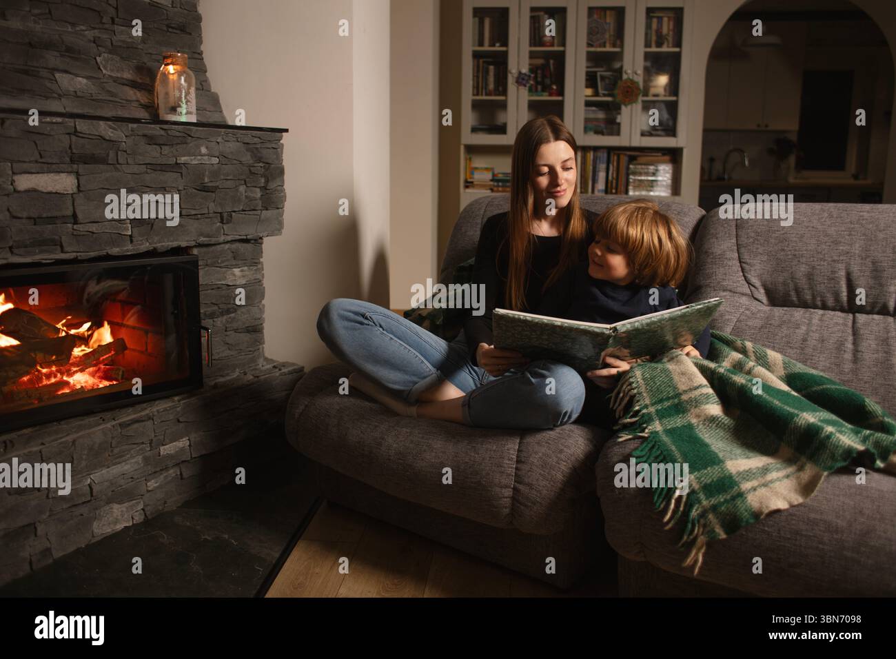Mère et fils lisent un livre sur un canapé près de la cheminée dans un intérieur confortable. Amour, confort et famille ensemble dans une maison rustique moderne. Banque D'Images