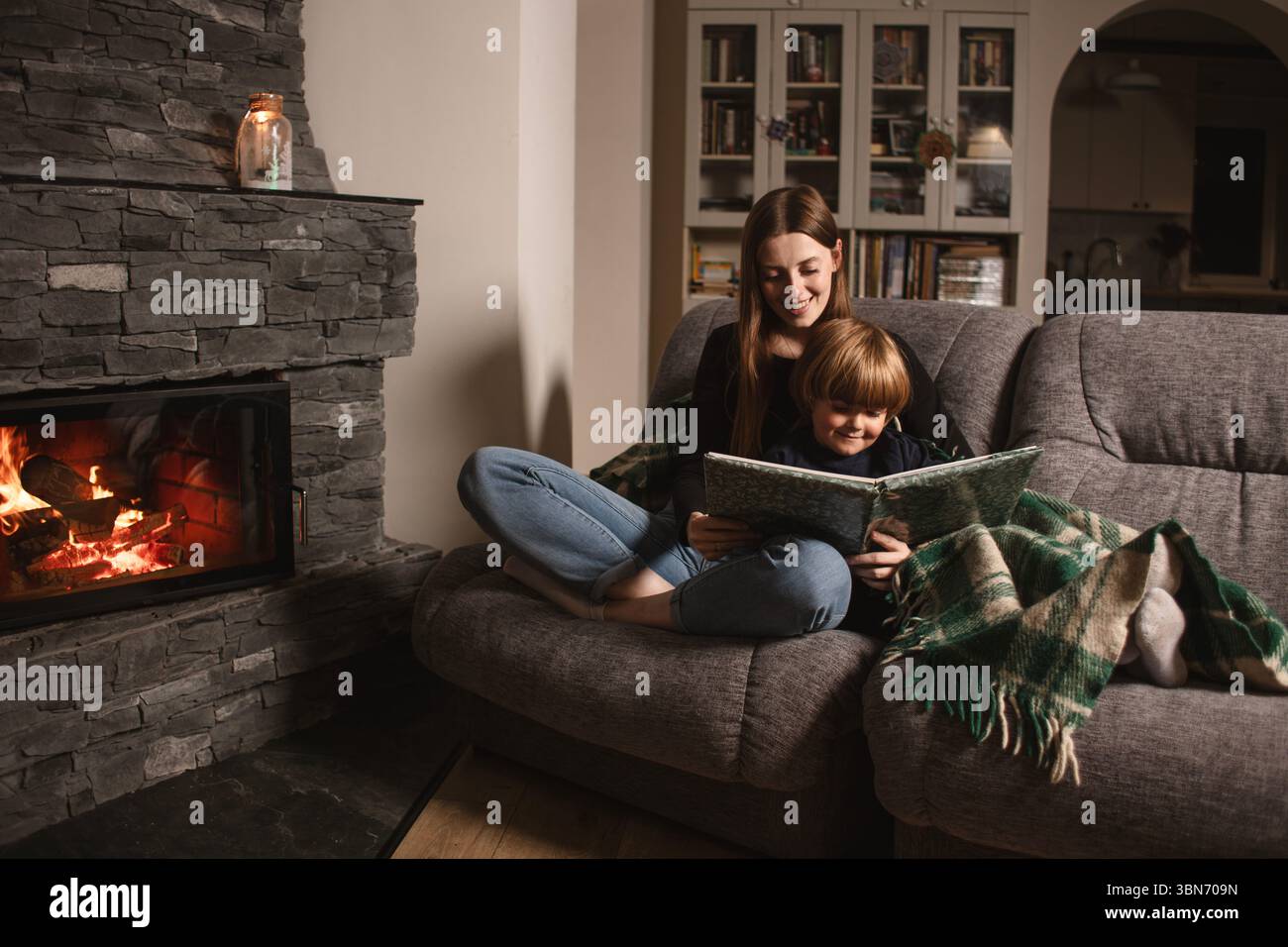 Mère et fils lisent un livre sur un canapé près de la cheminée dans un intérieur confortable. Amour, confort et famille ensemble dans une maison rustique moderne. Banque D'Images