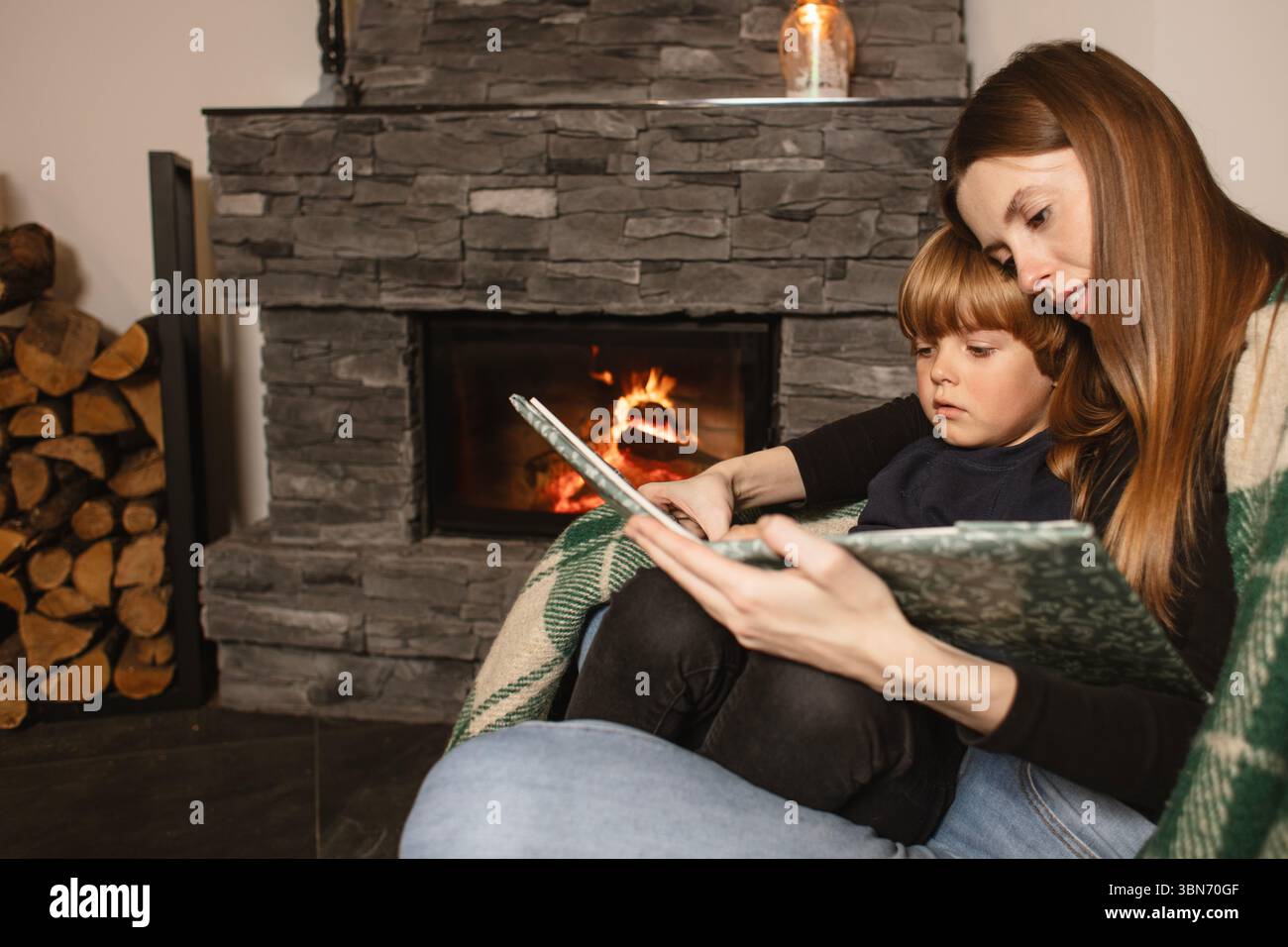 Mère et fils lisent un livre sur un canapé près de la cheminée dans un intérieur confortable. Amour, confort et famille ensemble dans une maison rustique moderne. Banque D'Images
