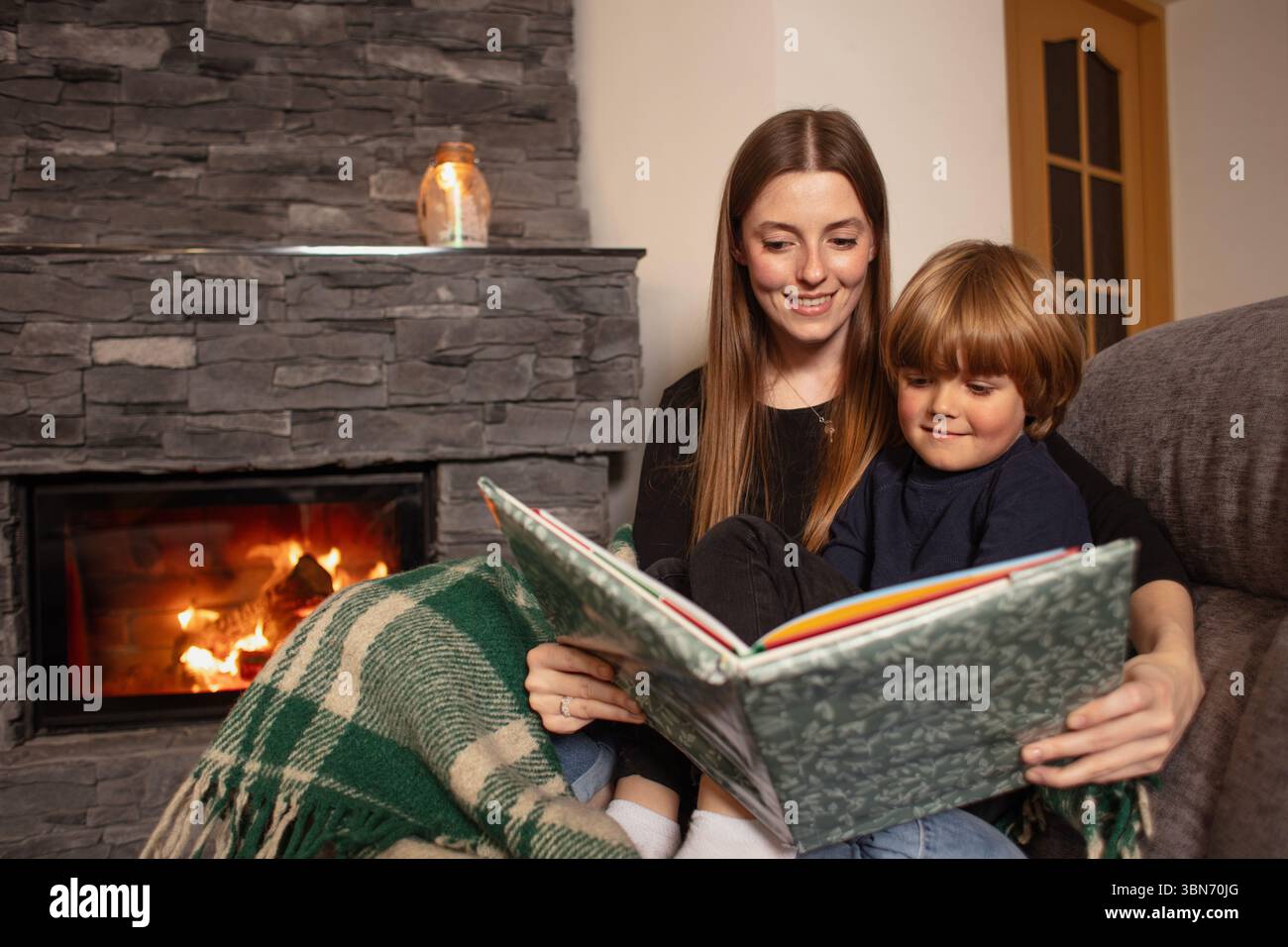 Mère et fils lisent un livre sur un canapé près de la cheminée dans un intérieur confortable. Amour, confort et famille ensemble dans une maison rustique moderne. Banque D'Images