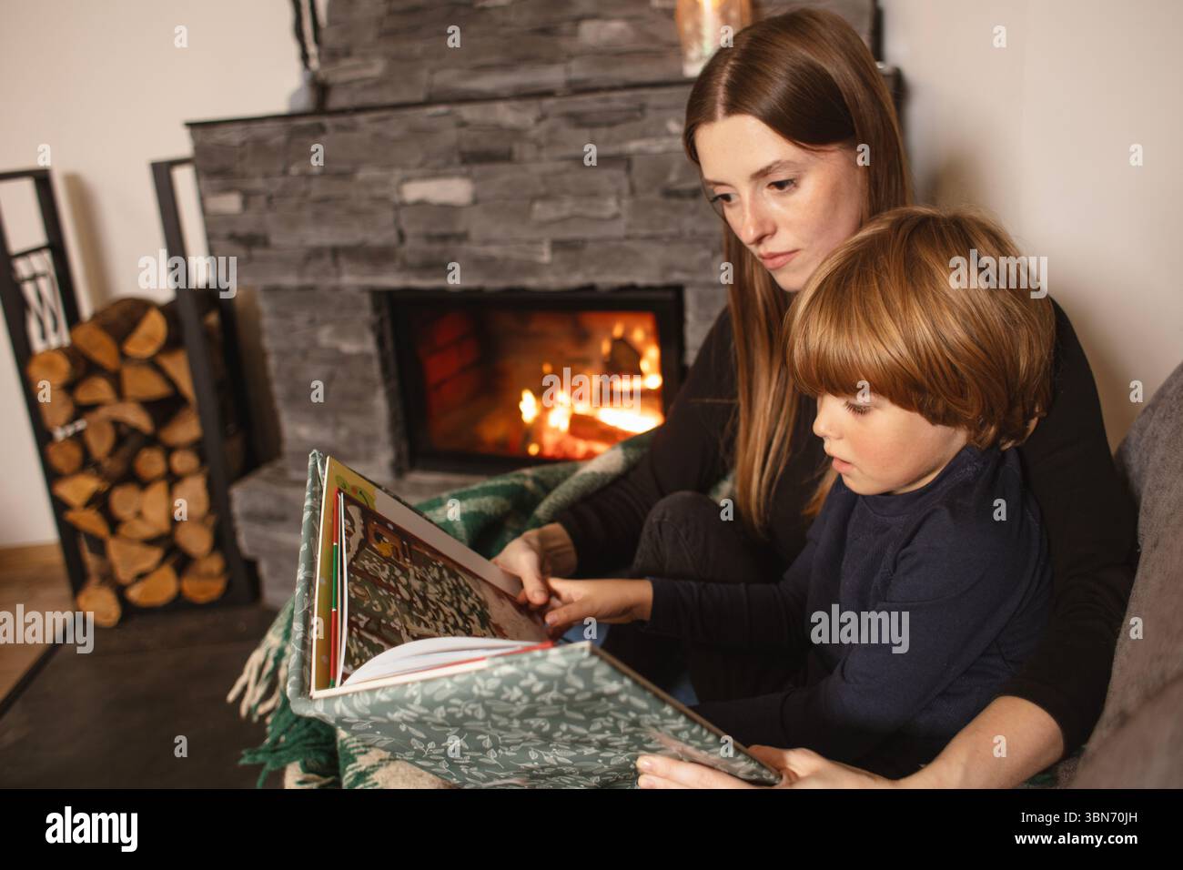 Mère et fils lisent un livre sur un canapé près de la cheminée dans un intérieur confortable. Amour, confort et famille ensemble dans une maison rustique moderne. Banque D'Images