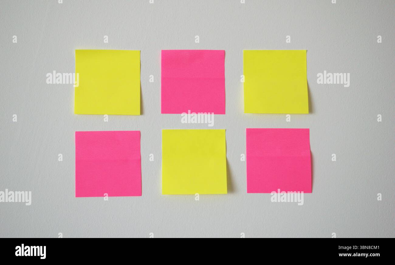Arrangement minimal et créatif de six notes autocollantes vierges en gros plan, trois jaunes et trois roses, soigneusement placées sur un mur blanc de couleur claire. Banque D'Images
