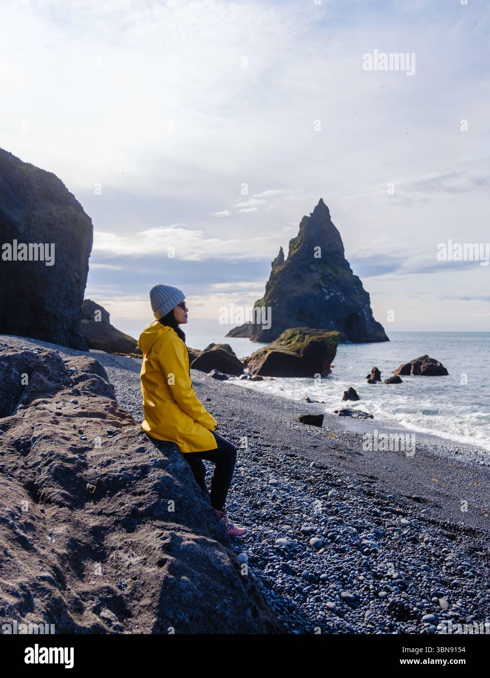 Une femme est captivée par la plage de Reynisfjara en Islande, connue pour son sable noir et ses colonnes de basalte imposantes. Une femme dans une veste de pluie jaune admire la beauté sauvage du paysage. Banque D'Images