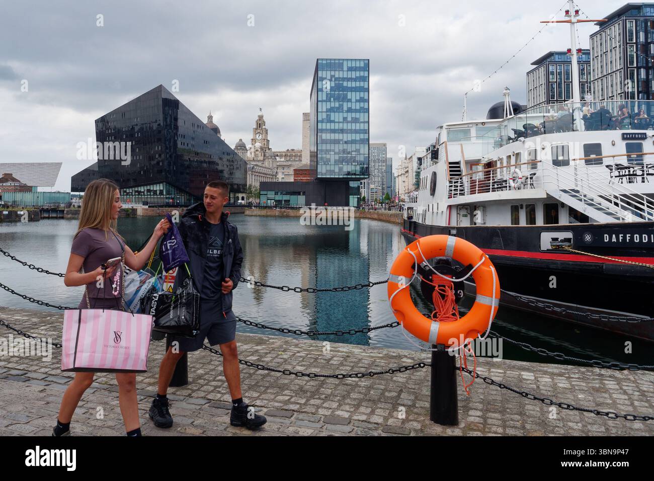 Jeune couple bronzé avec des sacs à provisions reflétant le style de vie des consommateurs marchent devant Canning Dock. Liverpool, Merseyside, Angleterre. 30 juin 2025 Banque D'Images