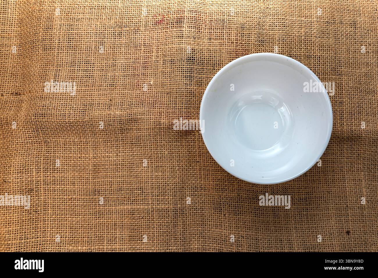 Bol en céramique blanche placé sur un fond de tissu rustique en toile de jute, vue de dessus. Idéal pour les maquettes de cuisine, la photographie culinaire, les blogs de recettes et la culine naturelle Banque D'Images