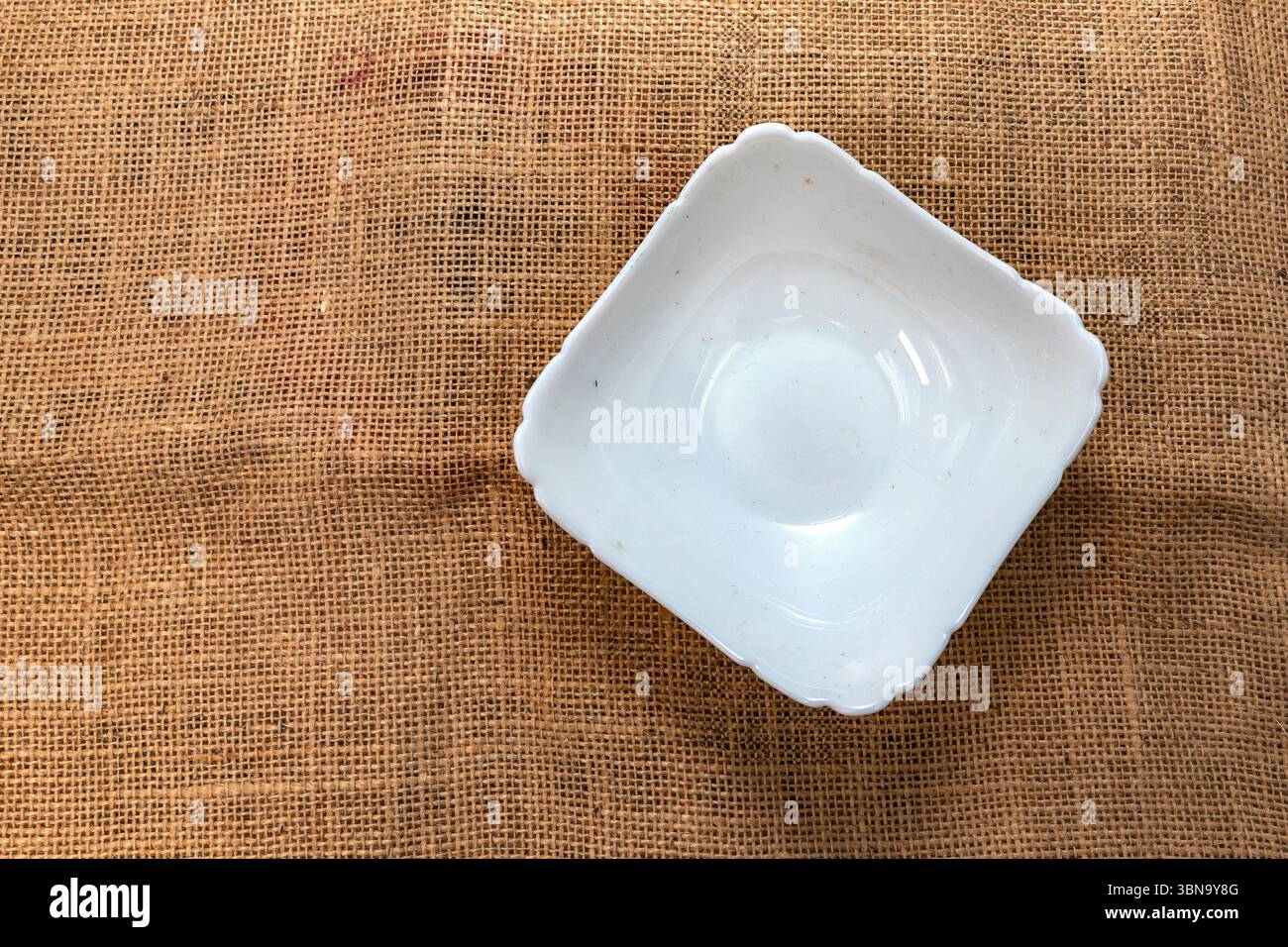Bol en céramique blanche placé sur un fond de tissu rustique en toile de jute, vue de dessus. Idéal pour les maquettes de cuisine, la photographie culinaire, les blogs de recettes et la culine naturelle Banque D'Images