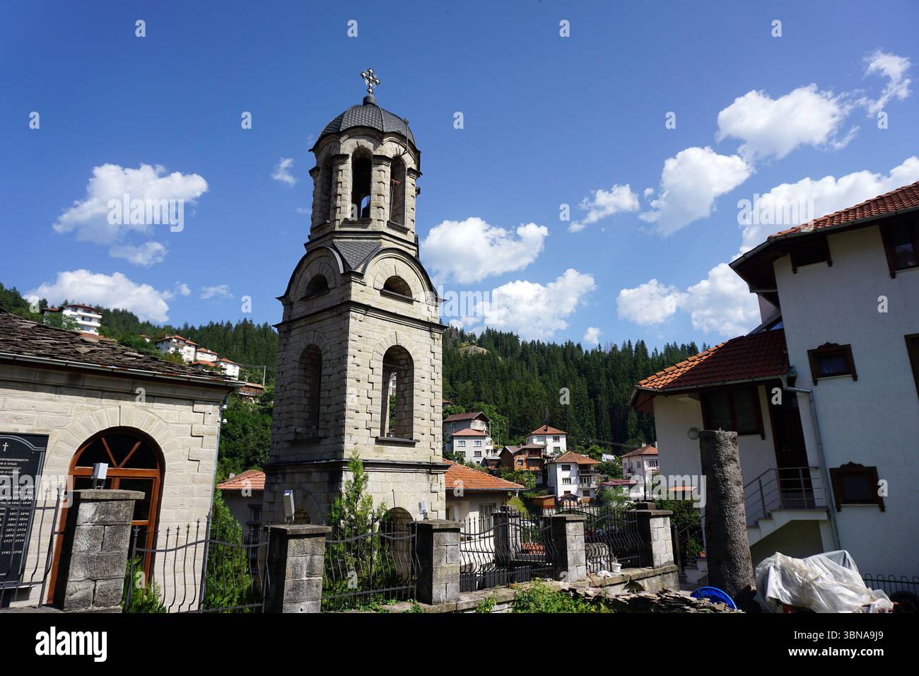 Momchilovtsi, un village dans les Rhodopes centraux en Bulgarie, Une scène d'église sereine. L'église, construite en pierre de couleur claire, est haute avec un clocher avec une croix à son sommet. Le clocher est orné de fenêtres cintrées et d'un toit gris foncé ou noir. L'église est entourée d'un mur de pierre avec une clôture métallique, et un petit bâtiment en pierre de couleur claire avec une porte en bois est visible à gauche. La région est nichée au milieu d'une forêt verdoyante, avec des maisons et des bâtiments de différentes tailles et couleurs, y compris des toits en tuiles blanches et rouges, dispersés dans le paysage. Le Banque D'Images