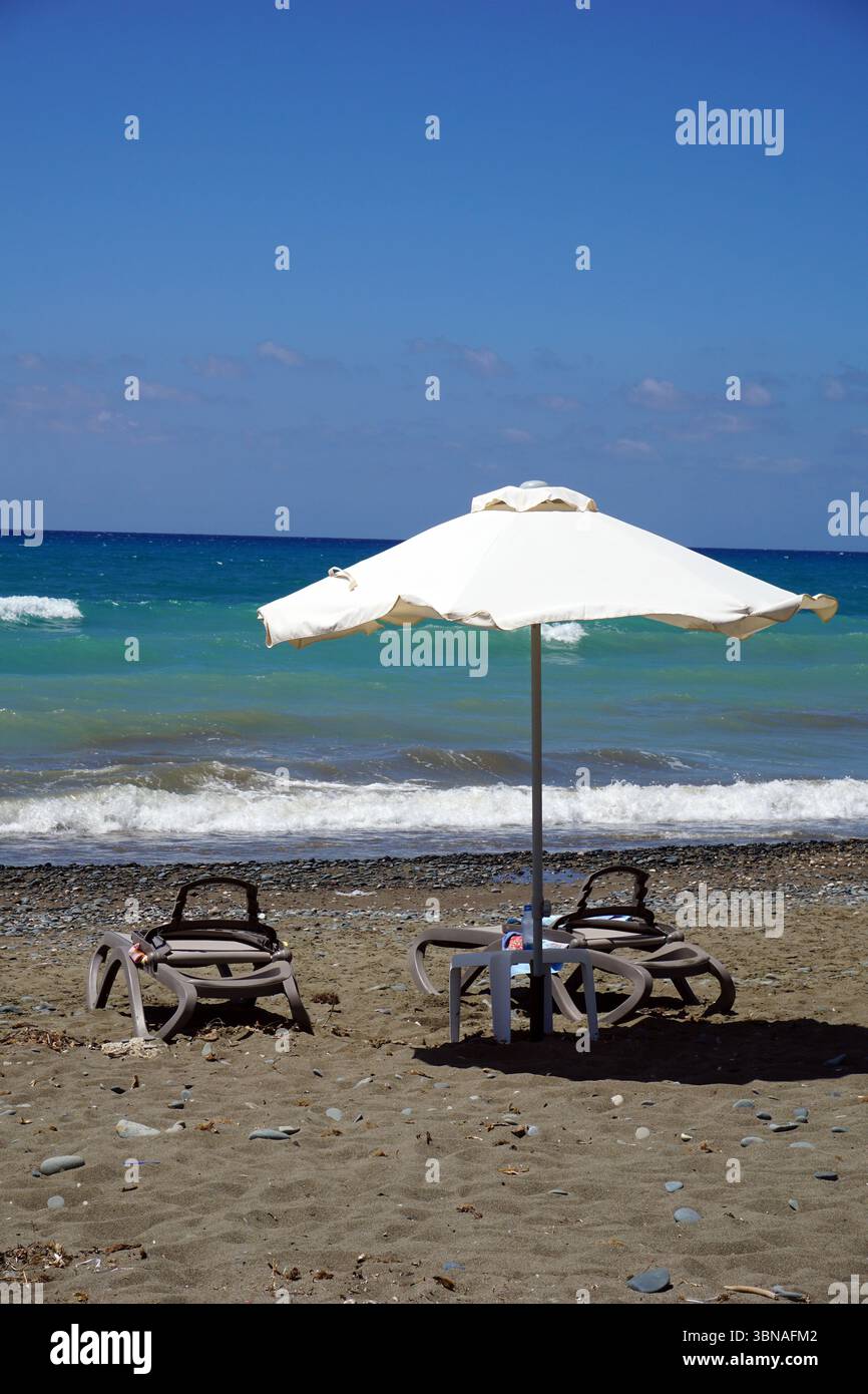 Cyprus Beach, Une scène de plage sereine. Au premier plan, un parapluie blanc est ouvert, offrant de l'ombre sur une petite table avec une nappe blanche. Deux chaises longues vides sont placées sous le parasol. La plage est couverte de sable brun clair, avec de petits cailloux dispersés à travers elle. L'océan, d'une couleur bleu-vert profond, s'étend en arrière-plan, avec de petites vagues qui se balancent doucement contre le rivage. Le ciel est d'un bleu clair et lumineux, et l'horizon est visible dans le Banque D'Images