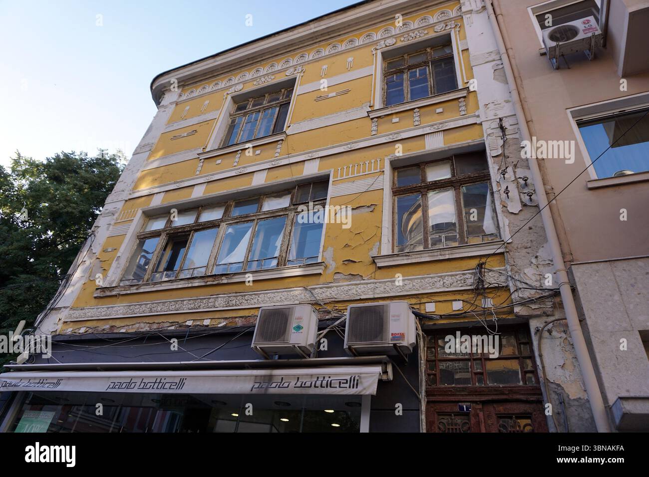 Plovdiv, la deuxième plus grande ville de Bulgarie, est l'une des plus anciennes villes d'Europe habitées en permanence, avec une histoire de plus de 8 000 ans. Niché le long de la rivière Maritsa., Un bâtiment jaune de trois étages avec des garnitures blanches et des détails ornés sur la façade. Le bâtiment montre des signes d'âge et de délabrement, avec de la peinture écaillée et certaines zones de plâtre manquant. Il a plusieurs fenêtres, dont certaines sont embarquées ou manquent des vitres. Un arbre est visible à gauche et une partie d'un autre bâtiment à droite. Une boutique avec un auvent blanc portant le nom 'pando botticelli' est visible sur le sol Banque D'Images