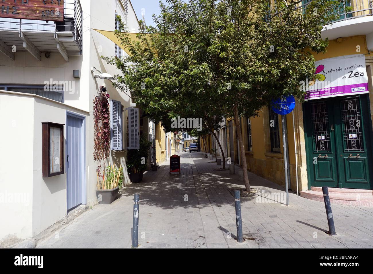 Une rue étroite bordée d'arbres dans un quartier de style méditerranéen. La rue est pavée de pierres grises et présente quelques arbres, dont un grand arbre aux feuilles vertes luxuriantes. Sur le côté droit de la rue, un bâtiment jaune avec une porte verte et un panneau qui indique "Zea Handmade Creations by Zeea" est visible. Un panneau bleu avec un symbole de vélo est également présent sur le côté droit de la rue. Le côté gauche de la rue présente un bâtiment blanc avec une porte bleue et une petite fenêtre de couleur foncée. Une petite poubelle rouge est placée près du centre de la rue. Un petit véhicule de couleur foncée est par Banque D'Images Une rue étroite bordée d'arbres dans un quartier de style méditerranéen. La rue est pavée de pierres grises et présente quelques arbres, dont un grand arbre aux feuilles vertes luxuriantes. Sur le côté droit de la rue, un bâtiment jaune avec une porte verte et un panneau qui indique "Zea Handmade Creations by Zeea" est visible. Un panneau bleu avec un symbole de vélo est également présent sur le côté droit de la rue. Le côté gauche de la rue présente un bâtiment blanc avec une porte bleue et une petite fenêtre de couleur foncée. Une petite poubelle rouge est placée près du centre de la rue. Un petit véhicule de couleur foncée est par Banque D'Images