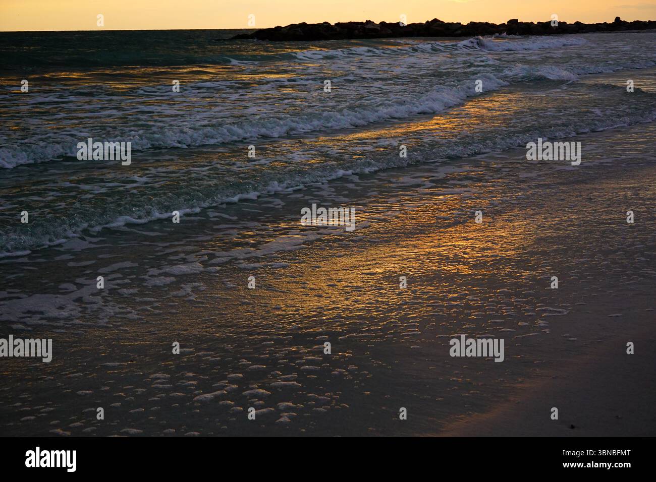 Lorsque le soleil se couche, la lumière dorée se reflète là où l'eau frappe le sable le long du rivage. Banque D'Images