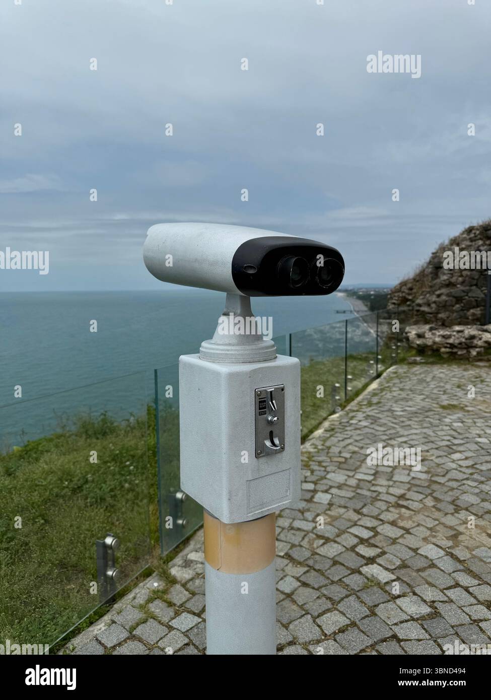 Cette image montre une visionneuse binoculaire publique avec une fente pour pièces de monnaie, installée sur un poteau dans une zone d'observation côtière. Il est positionné près d'un coffre-fort en verre Banque D'Images