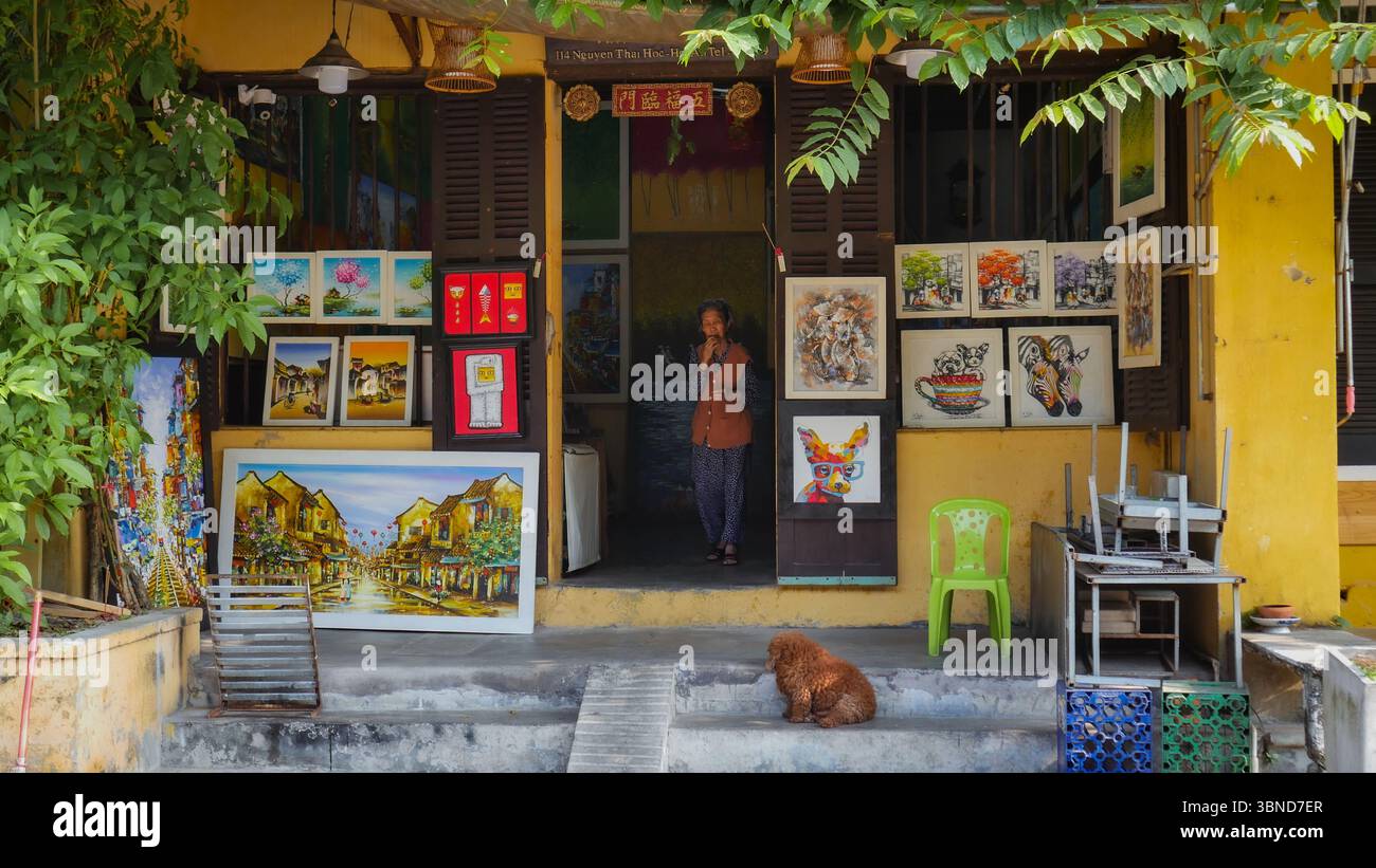 Galerie d'art colorée dans l'ancienne ville de Hoi an, Vietnam, avec femme et chien locaux devant le bâtiment traditionnel peint en jaune Banque D'Images