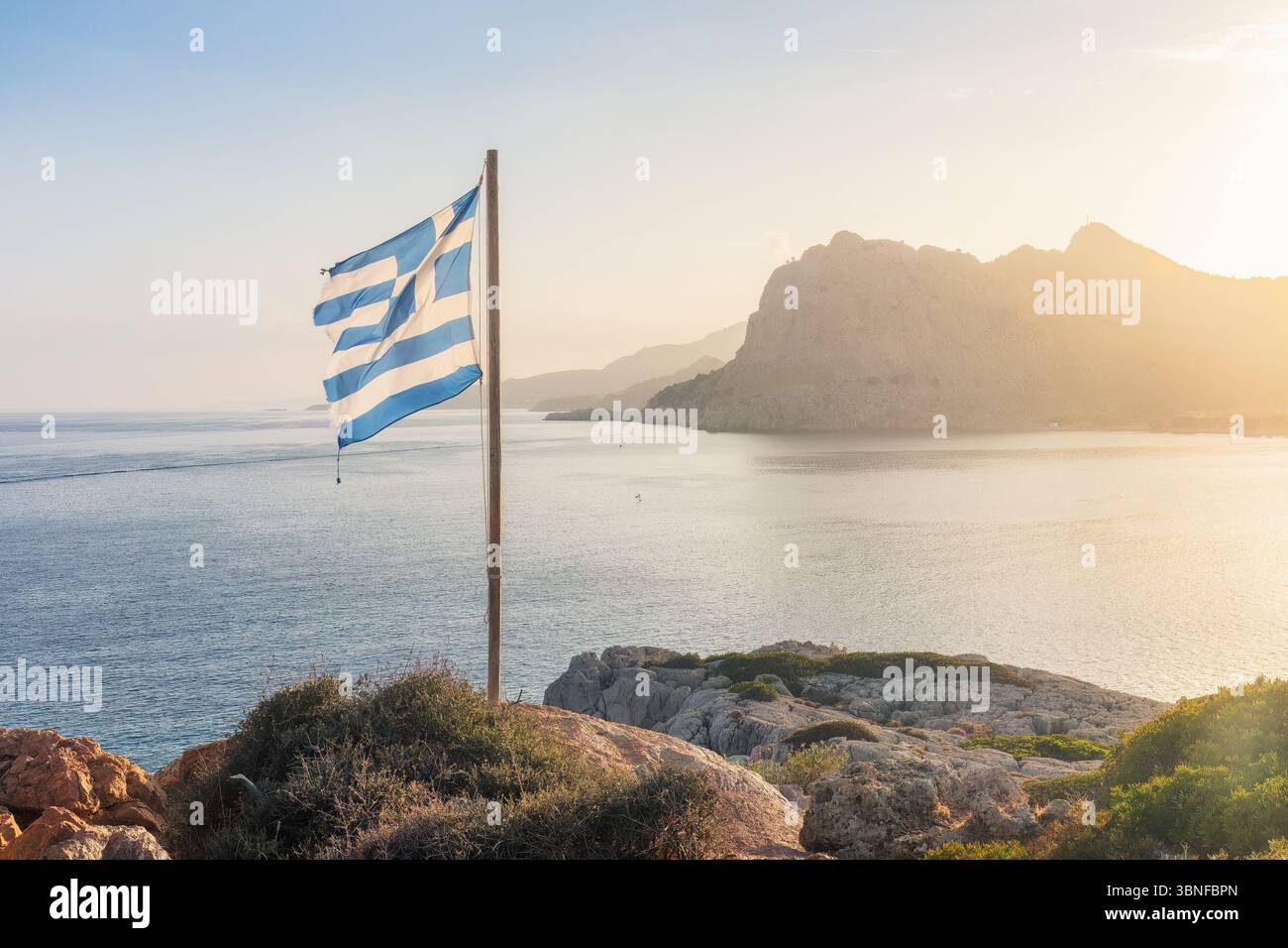 Falaises de bord de mer avec un drapeau flottant dans la brise pendant l'heure dorée, mettant en valeur les eaux turquoise et les douces teintes pastel. Grèce, île de Rhodes, Banque D'Images