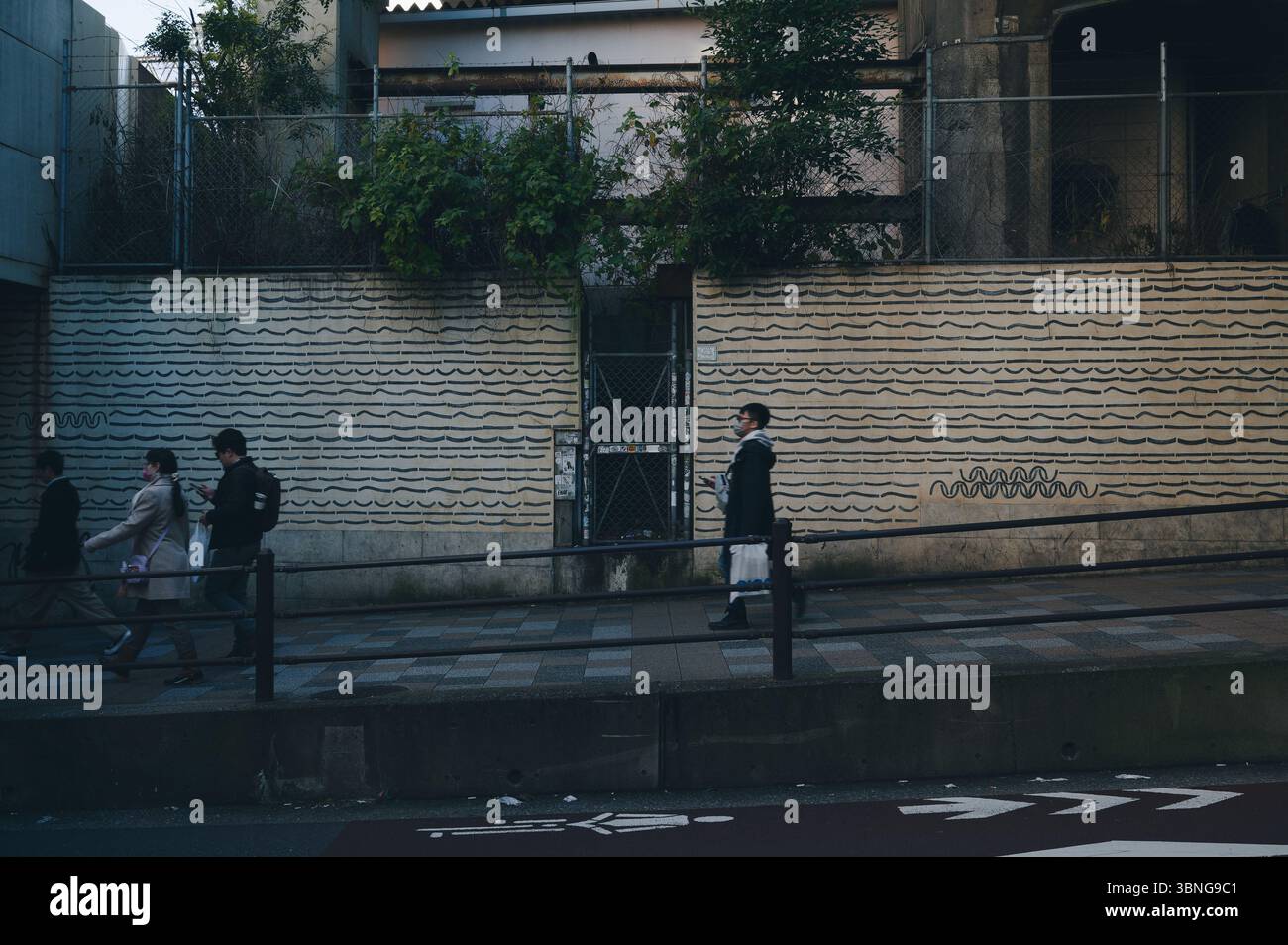 Les gens marchent devant un mur aux motifs minimalistes de vagues noires à Tokyo, encadrés par la verdure urbaine et les couleurs douces de l'hiver Banque D'Images